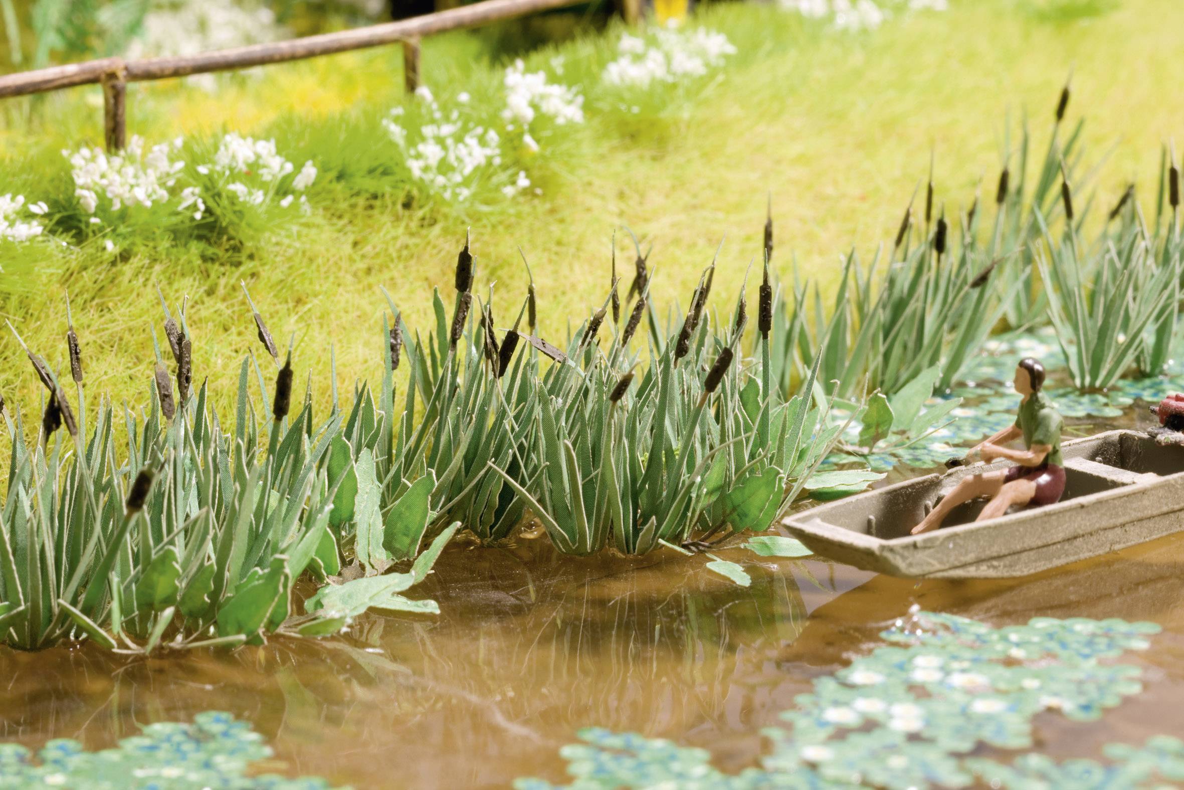 A miniature landscape depicts a person in a boat on a small river, surrounded by green plants and reeds along the riverbank.