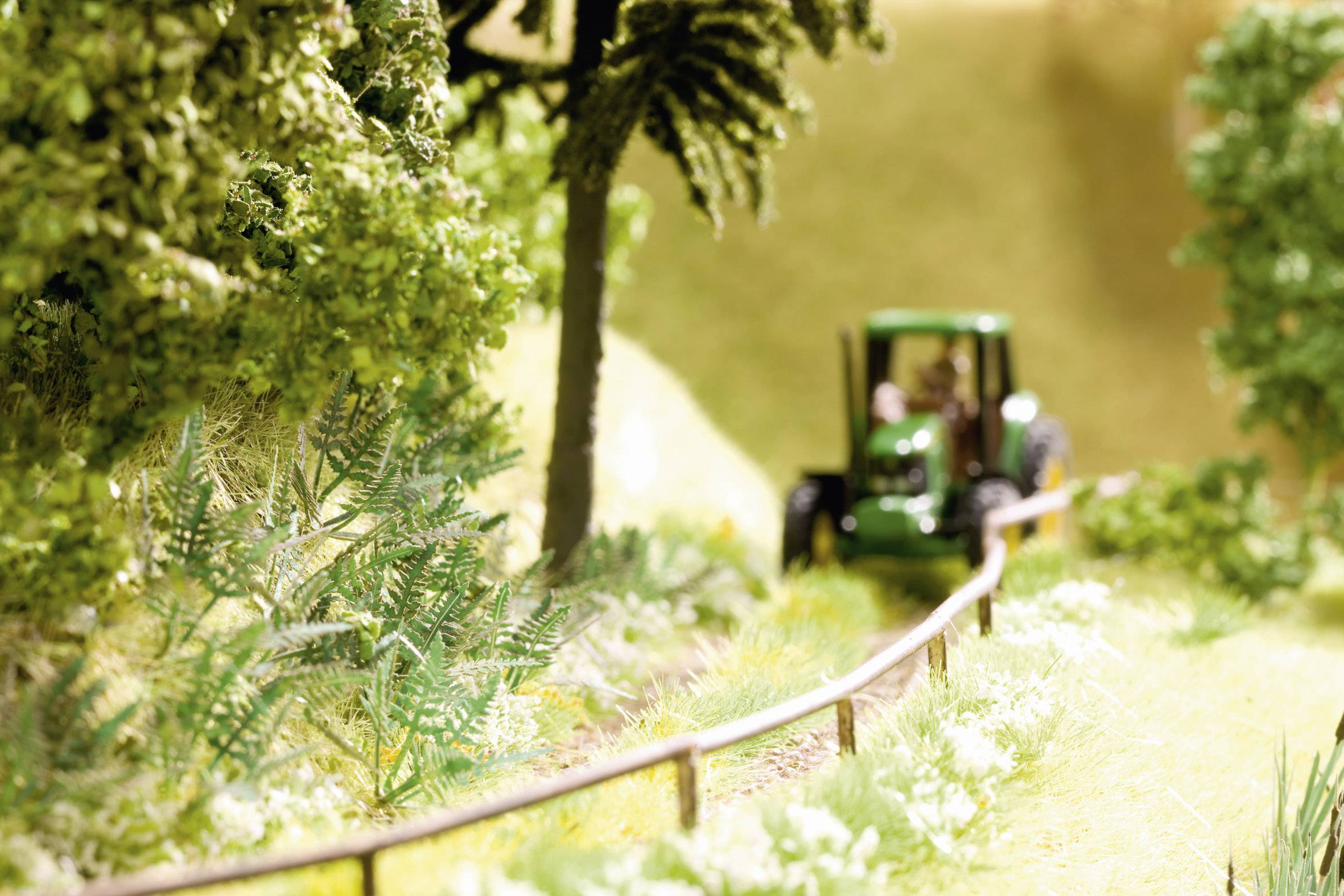 A small toy model of a green tractor drives along a country lane through lush green grass and past trees.