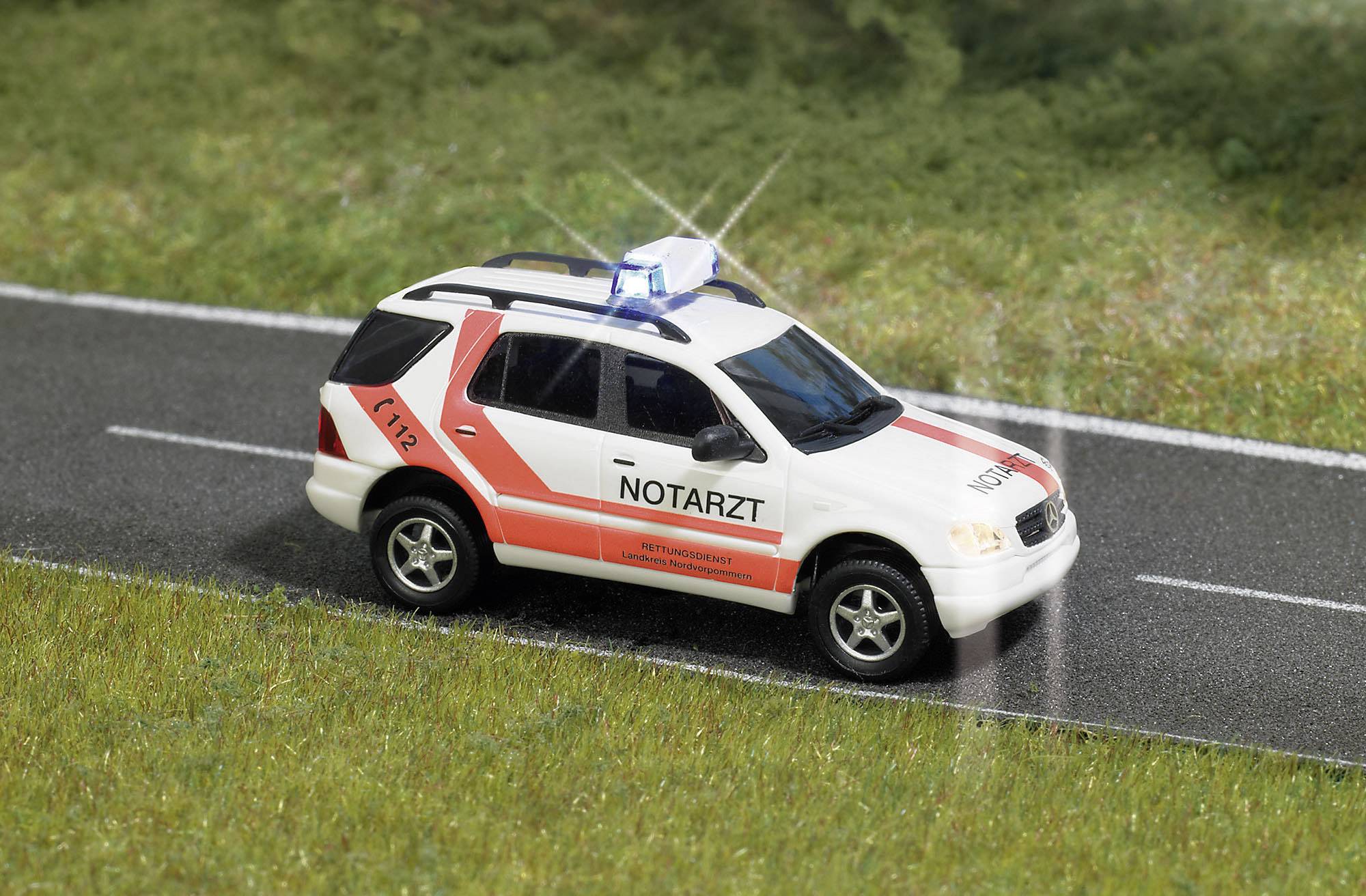 A white and red emergency medical response vehicle with its blue emergency lights activated is driving along a rural road.