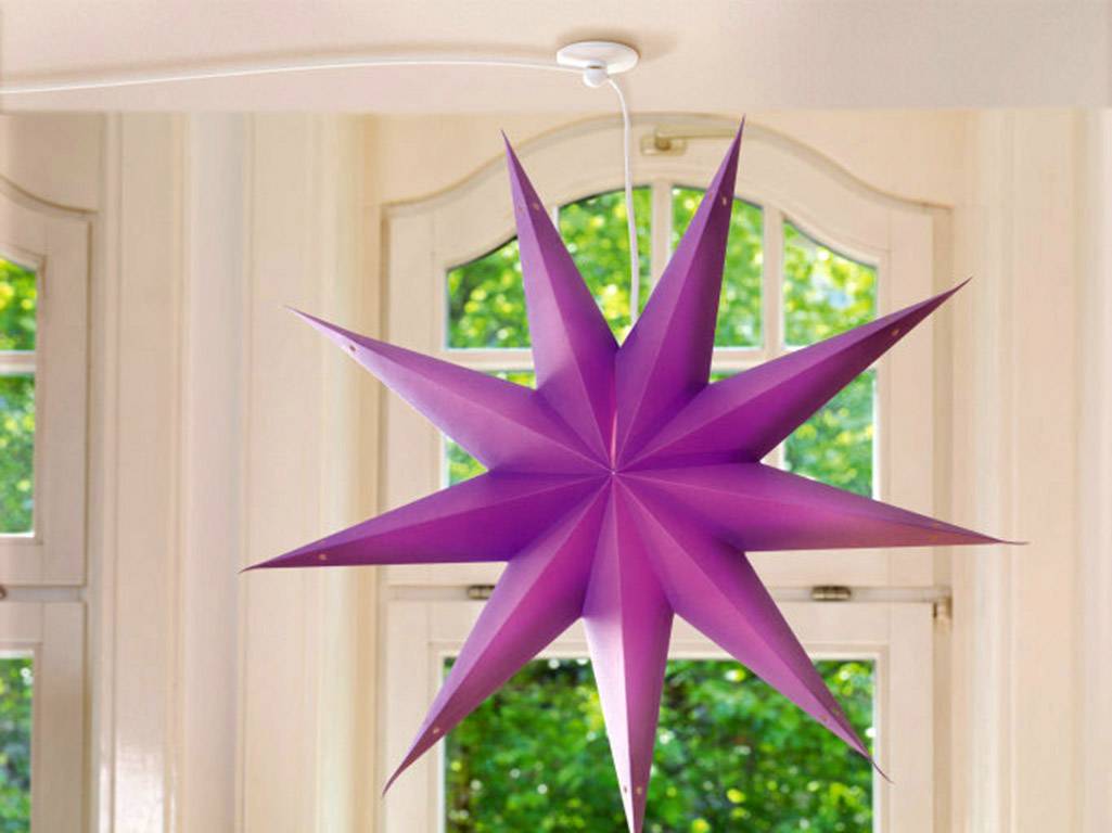 A large, purple paper star with eight points hangs from the ceiling in front of a bright window overlooking green trees.
