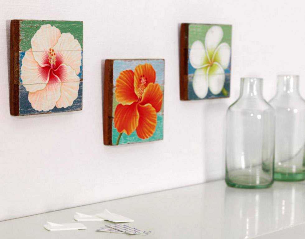 Three square pictures with floral motifs hang on a white wall. Next to them, two empty glass bottles stand on a smooth surface.
