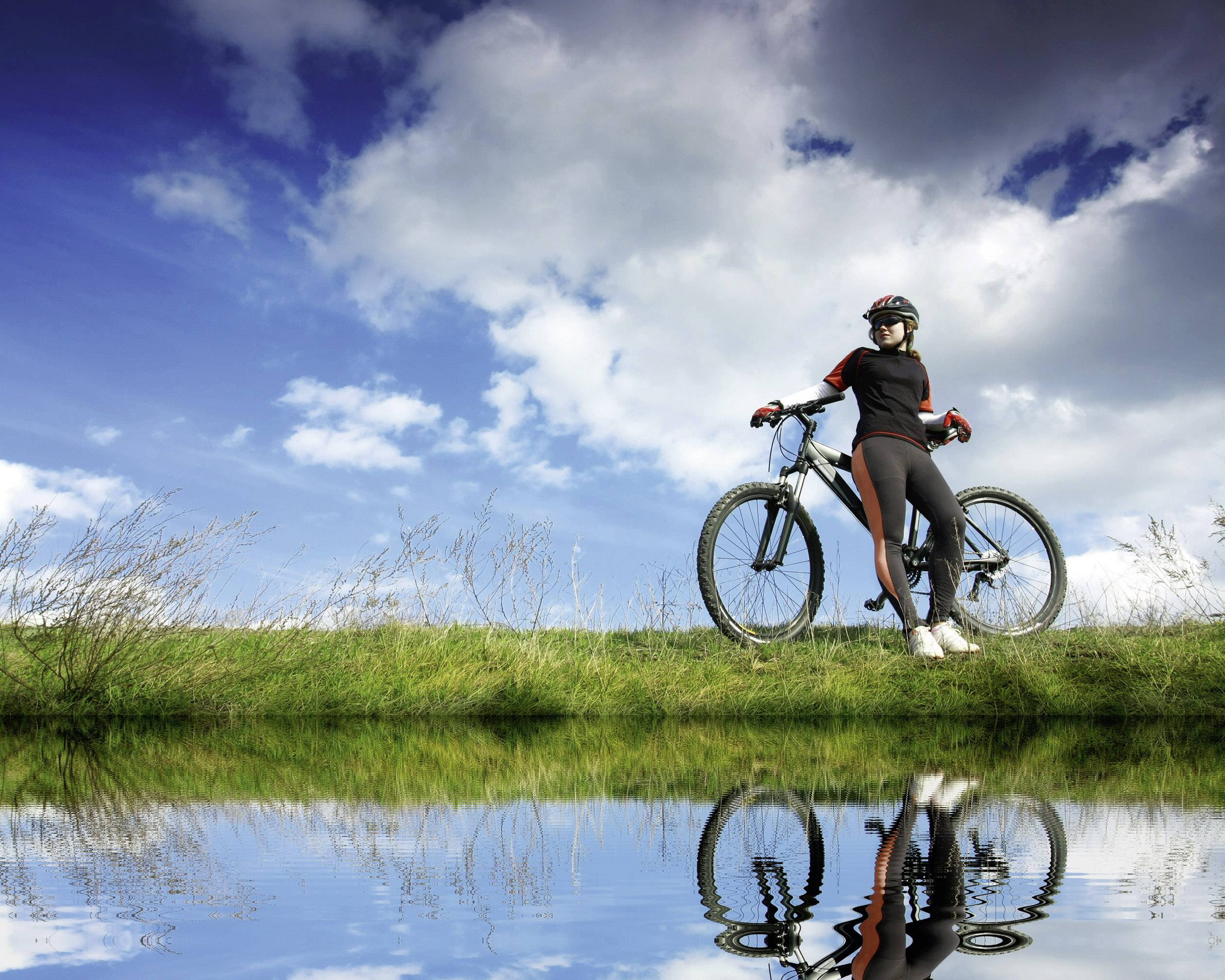 A person stands with their bicycle on the bank of a body of water beneath a blue sky with clouds. The image is reflected in the water.