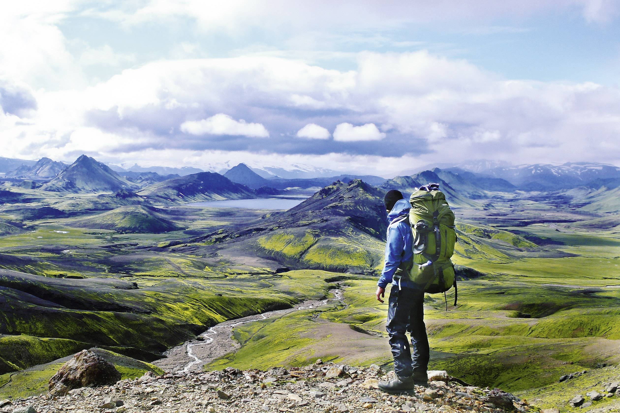 A person stands with a large rucksack on a hill, gazing out over a vast, green landscape with mountains and clouds on the horizon.