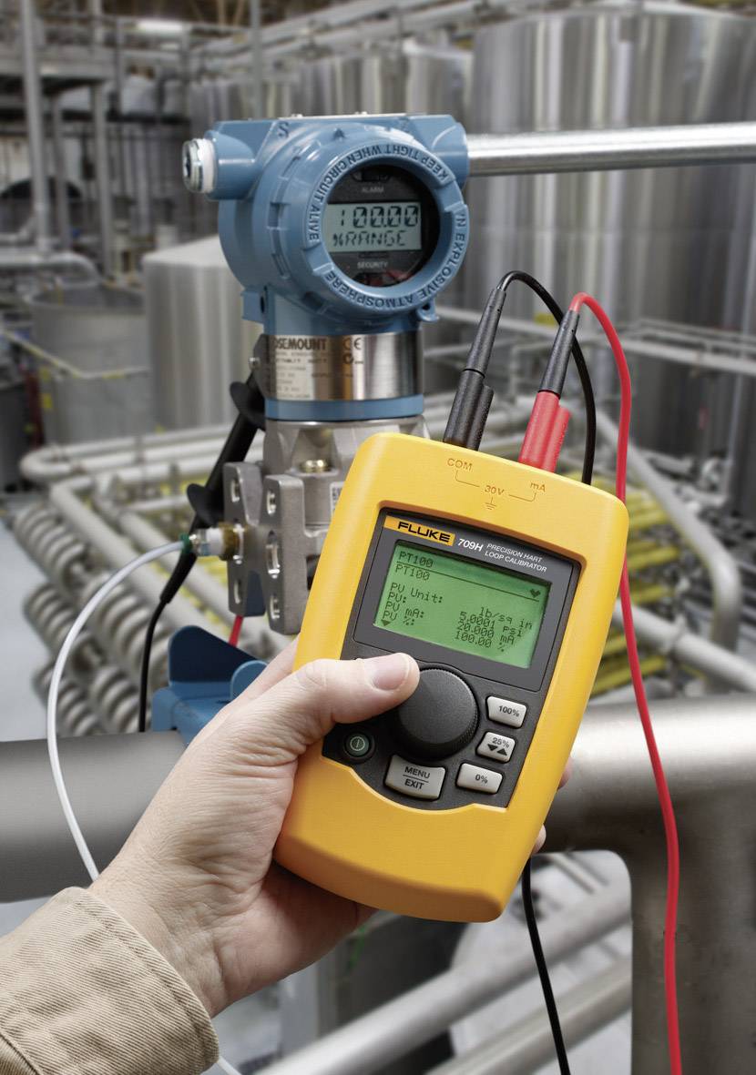A person is measuring the pressure of a blue industrial pressure sensor using a yellow digital multimeter, surrounded by metal pipes.