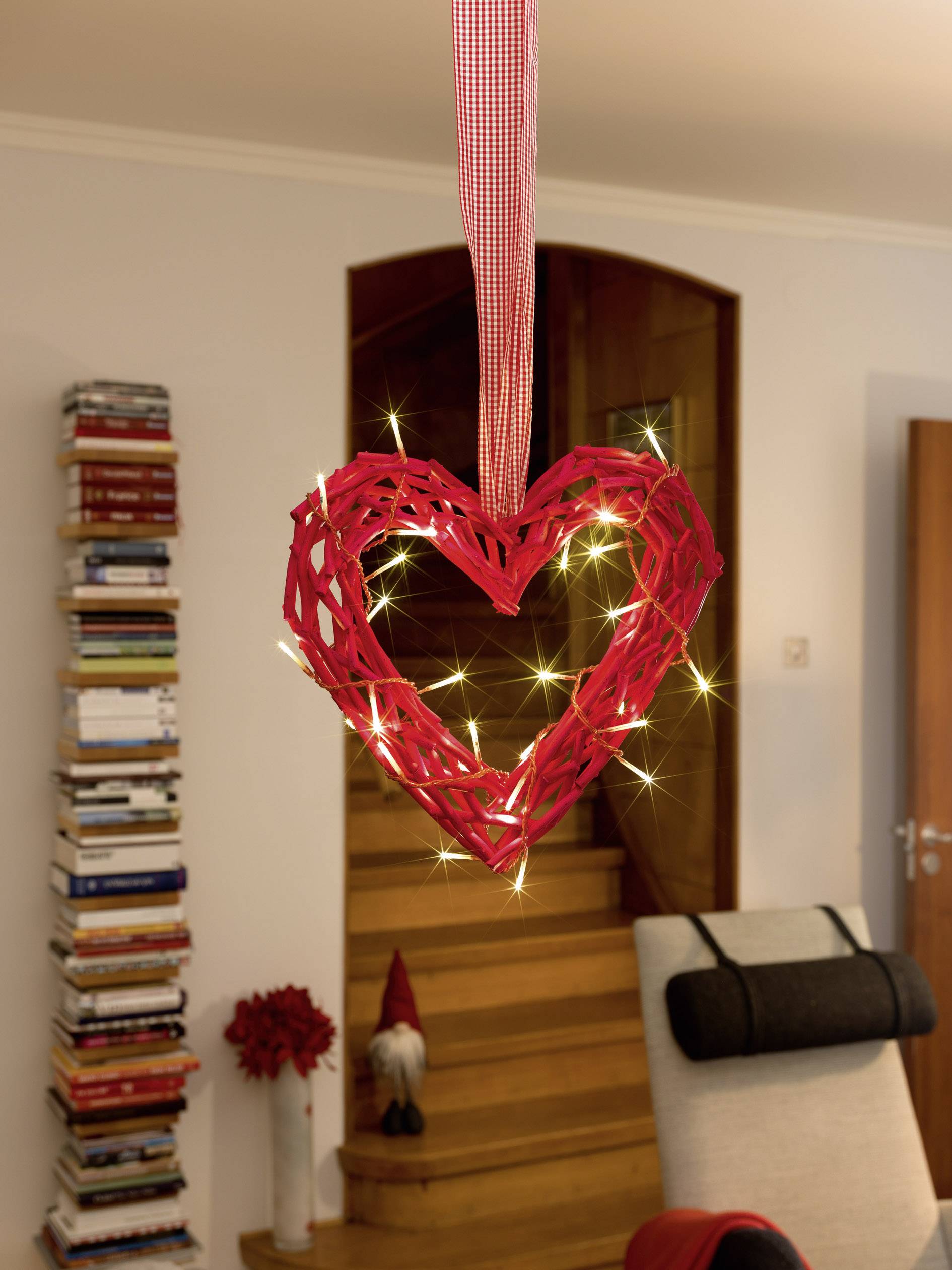 A braided red heart, illuminated by small lights, hangs in front of a wooden staircase. A stack of books and a gnome are in the background.