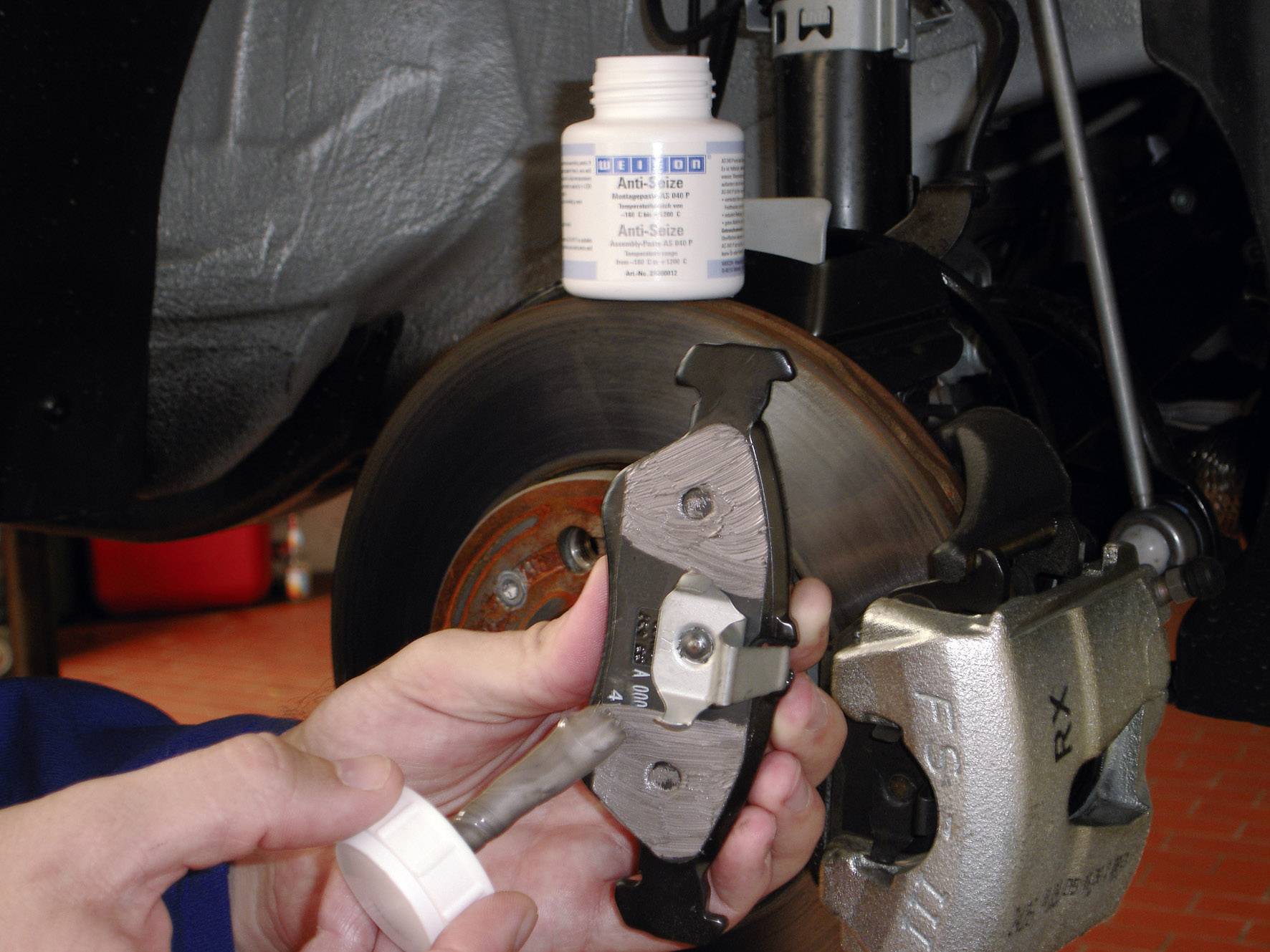 A person is applying paste to the back of a brake pad, in front of an open vehicle brake system. A container of 'anti-squeal' paste is placed on top.