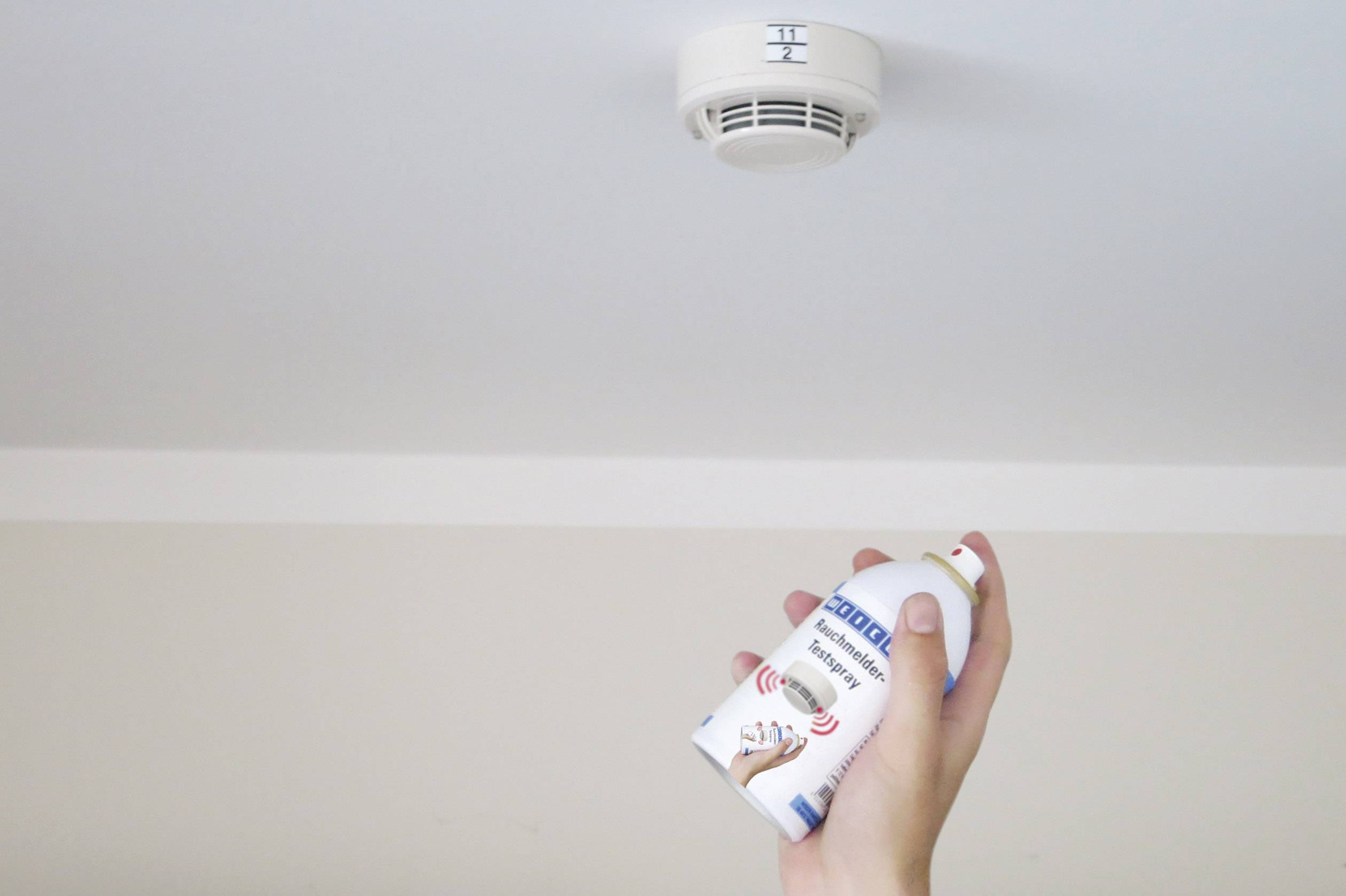 A hand is testing a smoke detector on the ceiling with a spray to ensure the device's functionality.