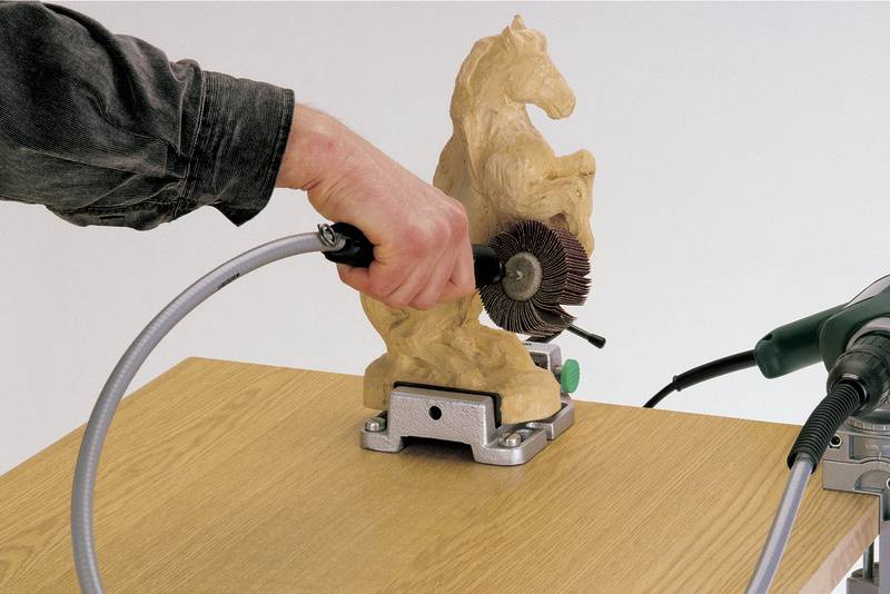A person is working on a wooden horse sculpture with a rotating grinding machine on a wooden table.