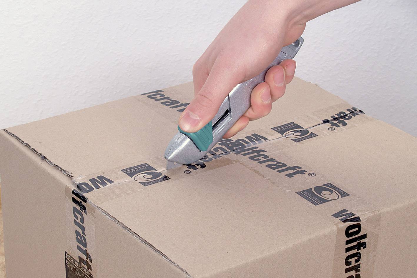 A hand opens a cardboard box sealed with tape printed with 'wolfcraft' using a utility knife.