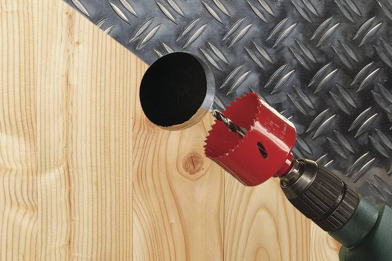 A drill with a red hole saw is boring through a piece of wood, with diamond-patterned metal visible in the background.