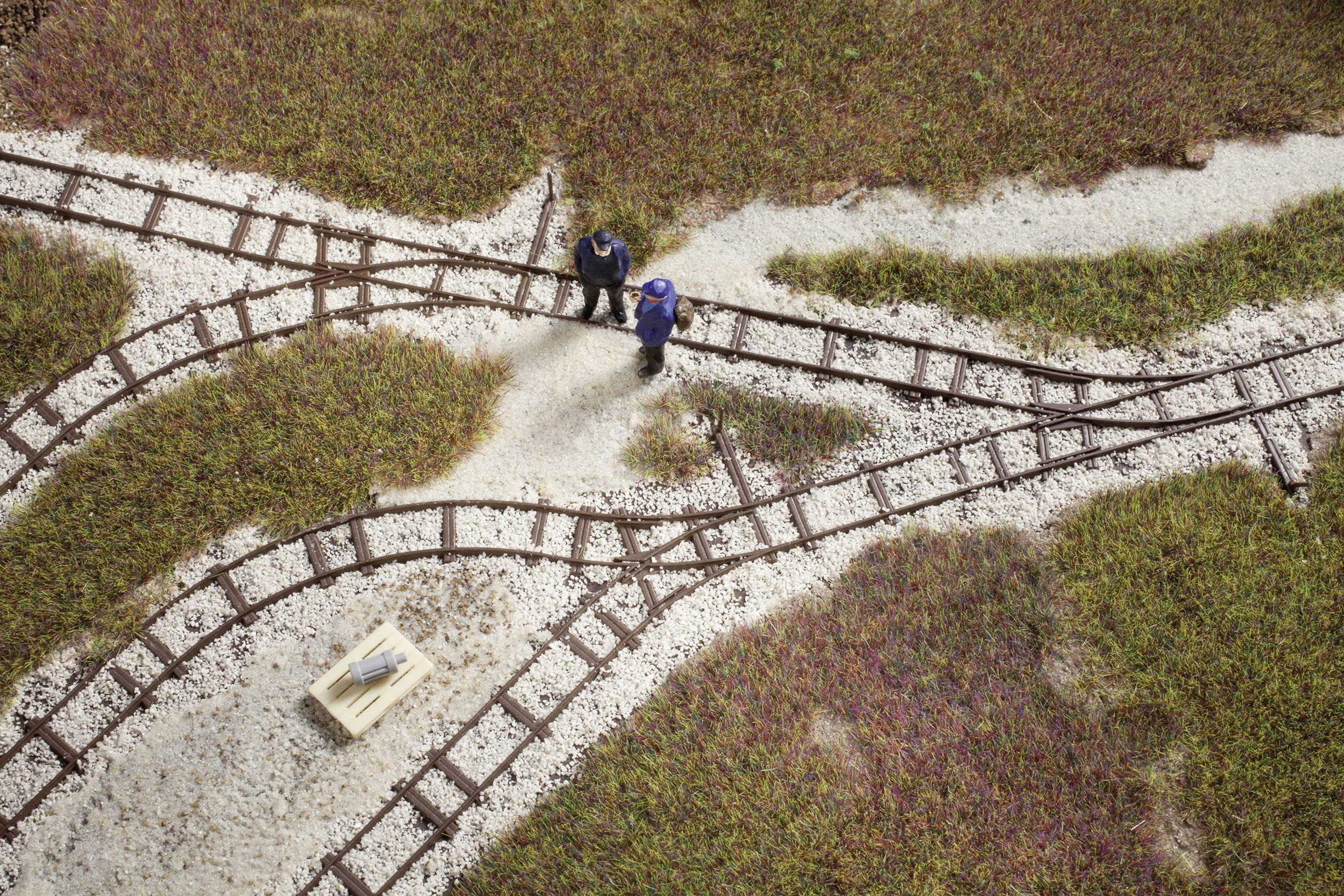Two people are standing on a model railway layout with points and tracks, surrounded by grassy areas. A cardboard box is nearby.