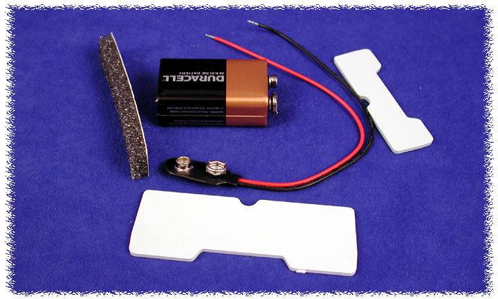 A 9-volt battery with a battery clip and wires lies beside two rectangular, white plastic components and a thin foam strip on a blue surface.