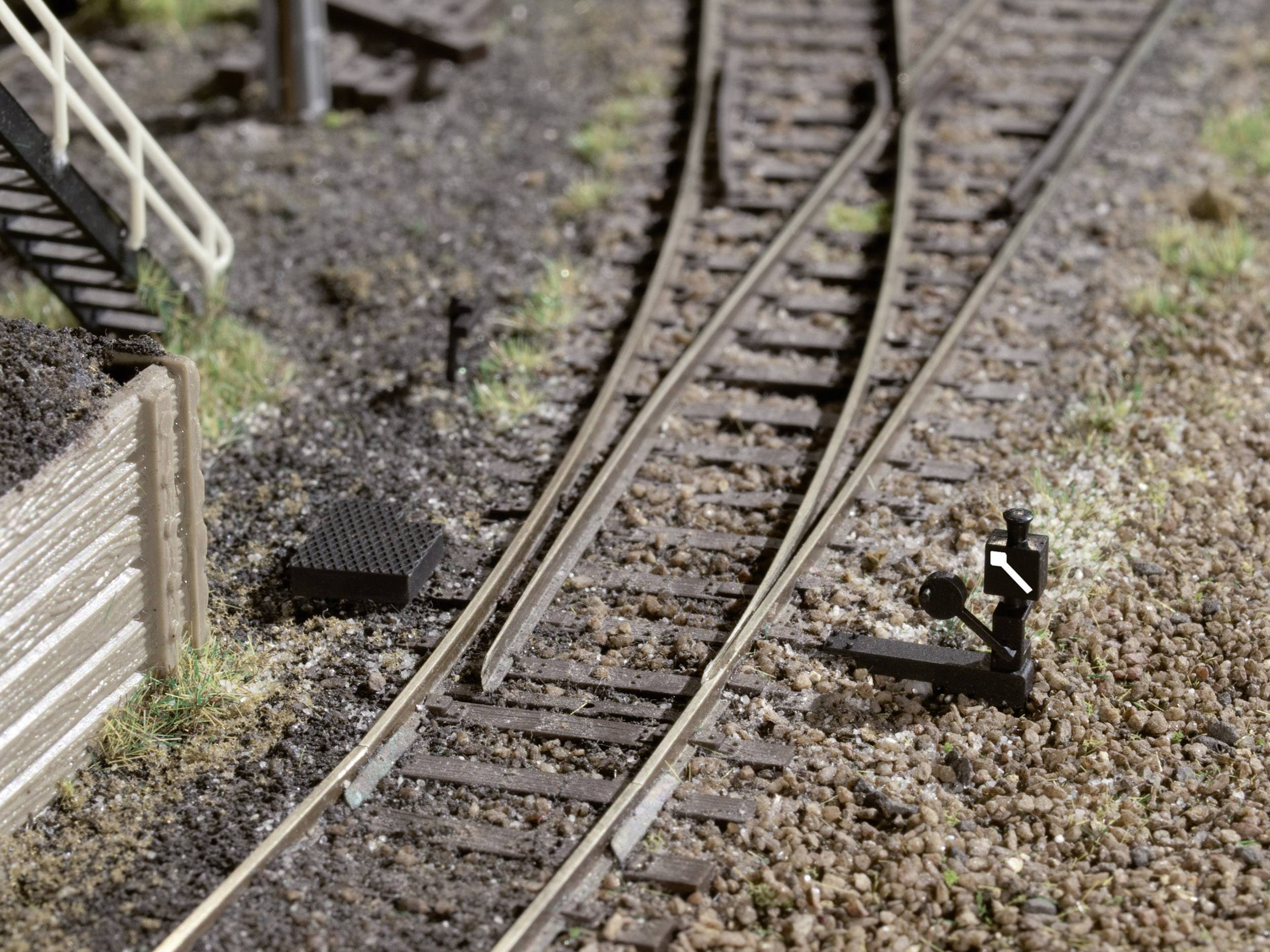 A railway model shows tracks with a points lever on the right and a wooden barrier on the left. There are grassy patches along the ballast beds.