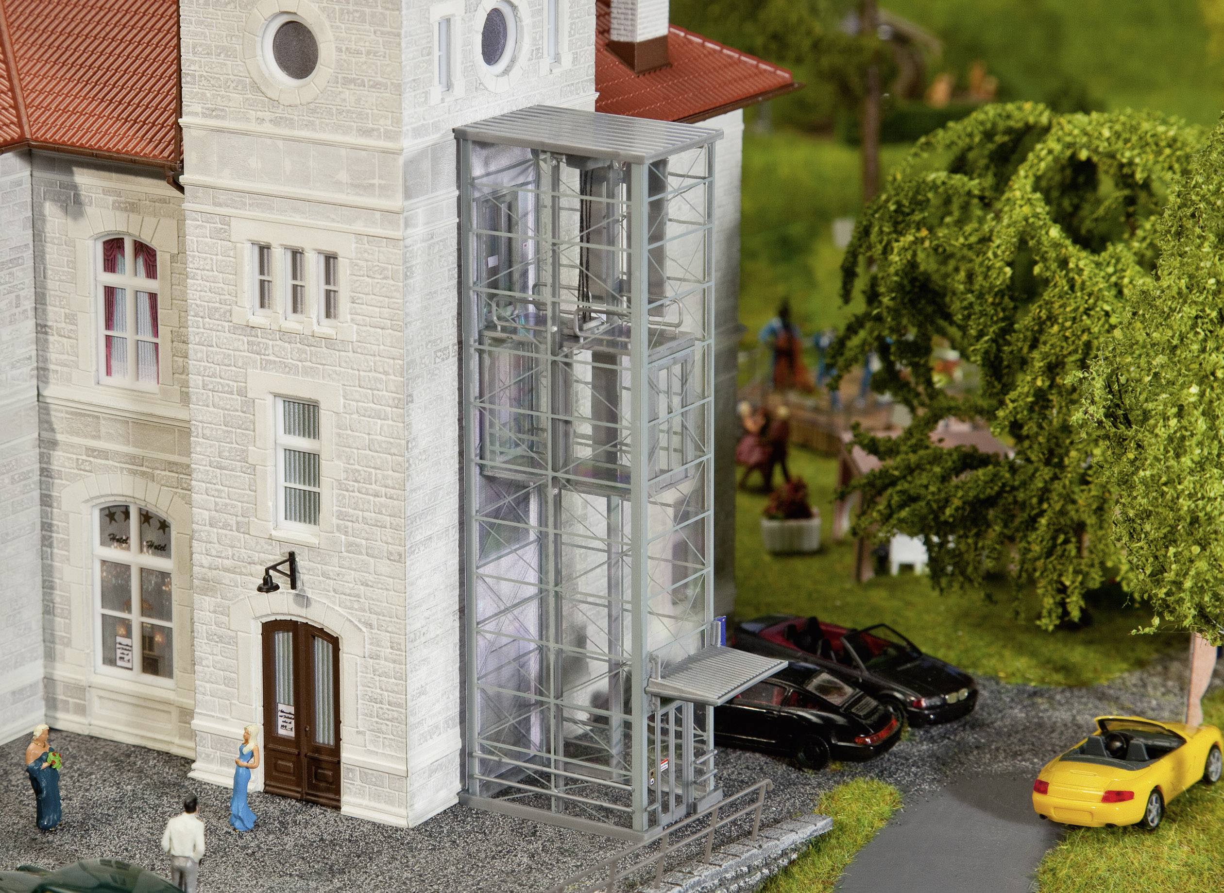 A miniature building with a glass lift, surrounded by trees and parked cars. People are standing and chatting in front of the building.