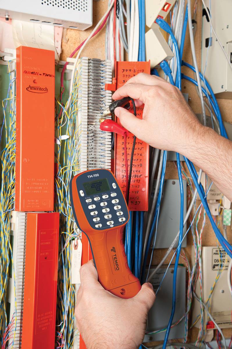 A person is testing a telecommunications distribution panel with a cable testing device. Various cables are visible around the panel.