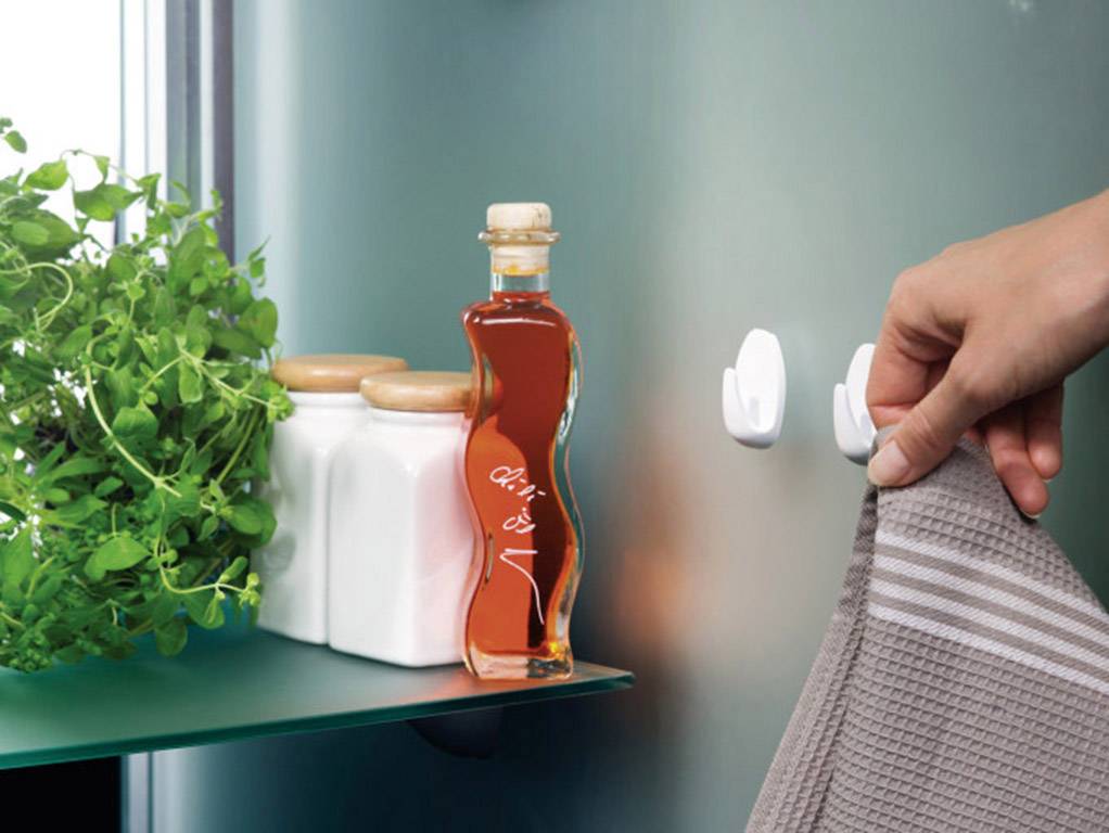 A hand hangs a grey towel on a hook. Next to it, herb pots and a bottle of oil are standing on a shelf.
