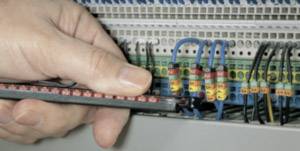 A hand holds a testing device and checks electrical connections in a control cabinet with wired connections.