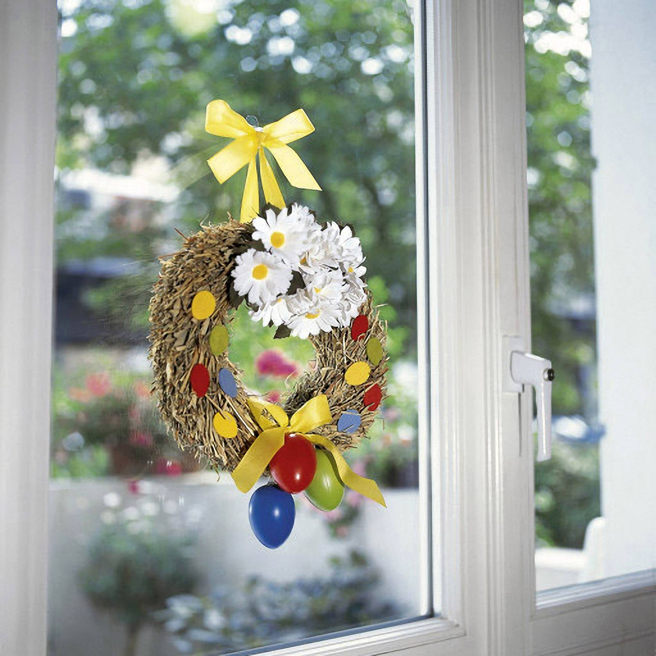 A colourful Easter wreath with yellow ribbons, white flowers, and colourful eggs hangs in a window. Outside, blurred trees can be seen.