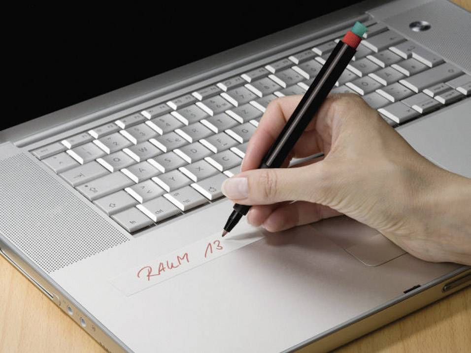 A hand writes 'ROOM 13' on a piece of tape stuck to a laptop using a pen.