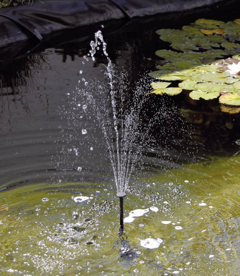 A small fountain in a garden pond, surrounded by floating leaves, creates bubbling water jets.