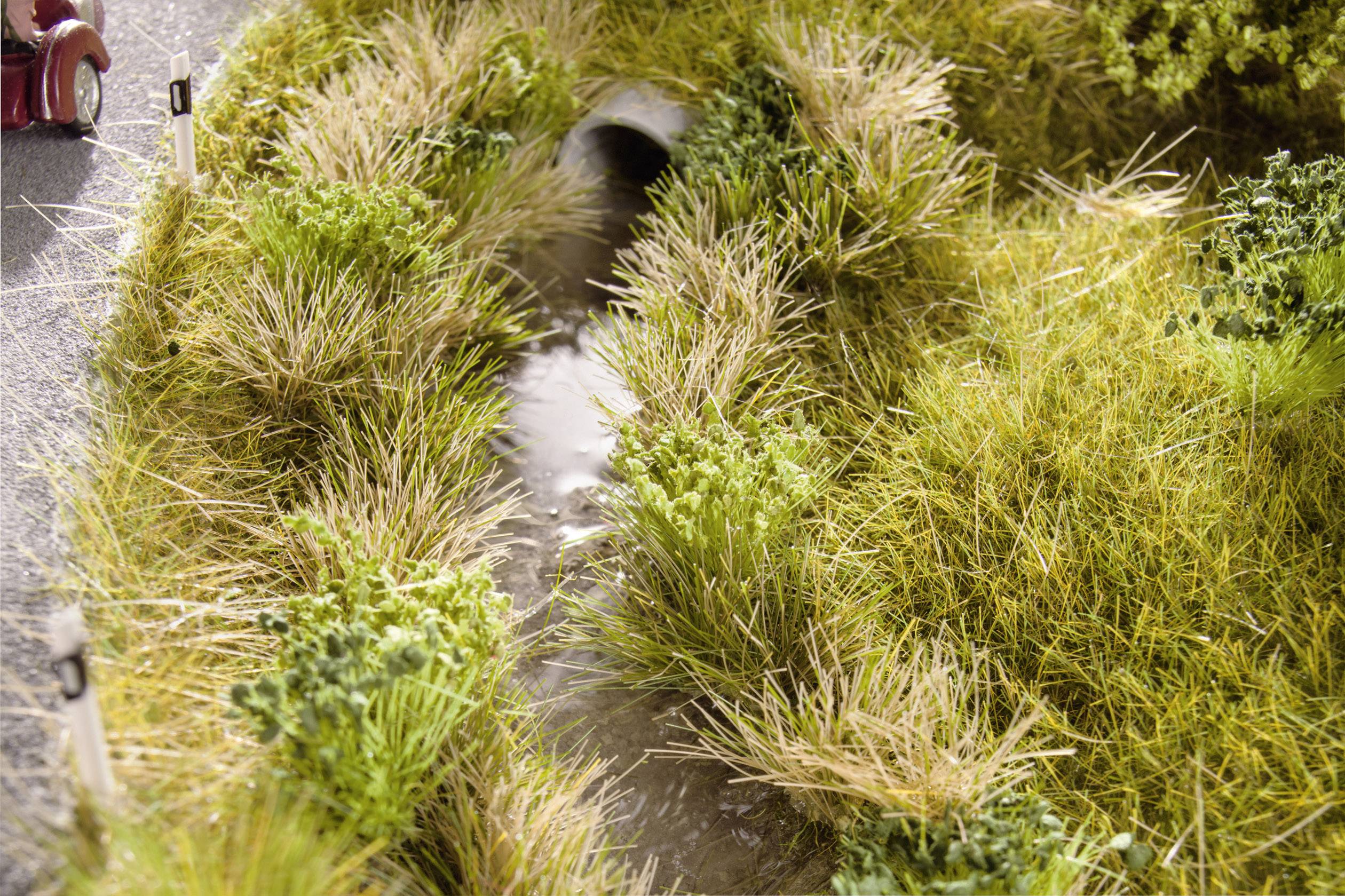 'A small, overgrown ditch beside a road, through which a narrow stream flows. Vegetation dominates the scene.'<br><br>Note: The translation is already in British English, so no significant changes were needed.