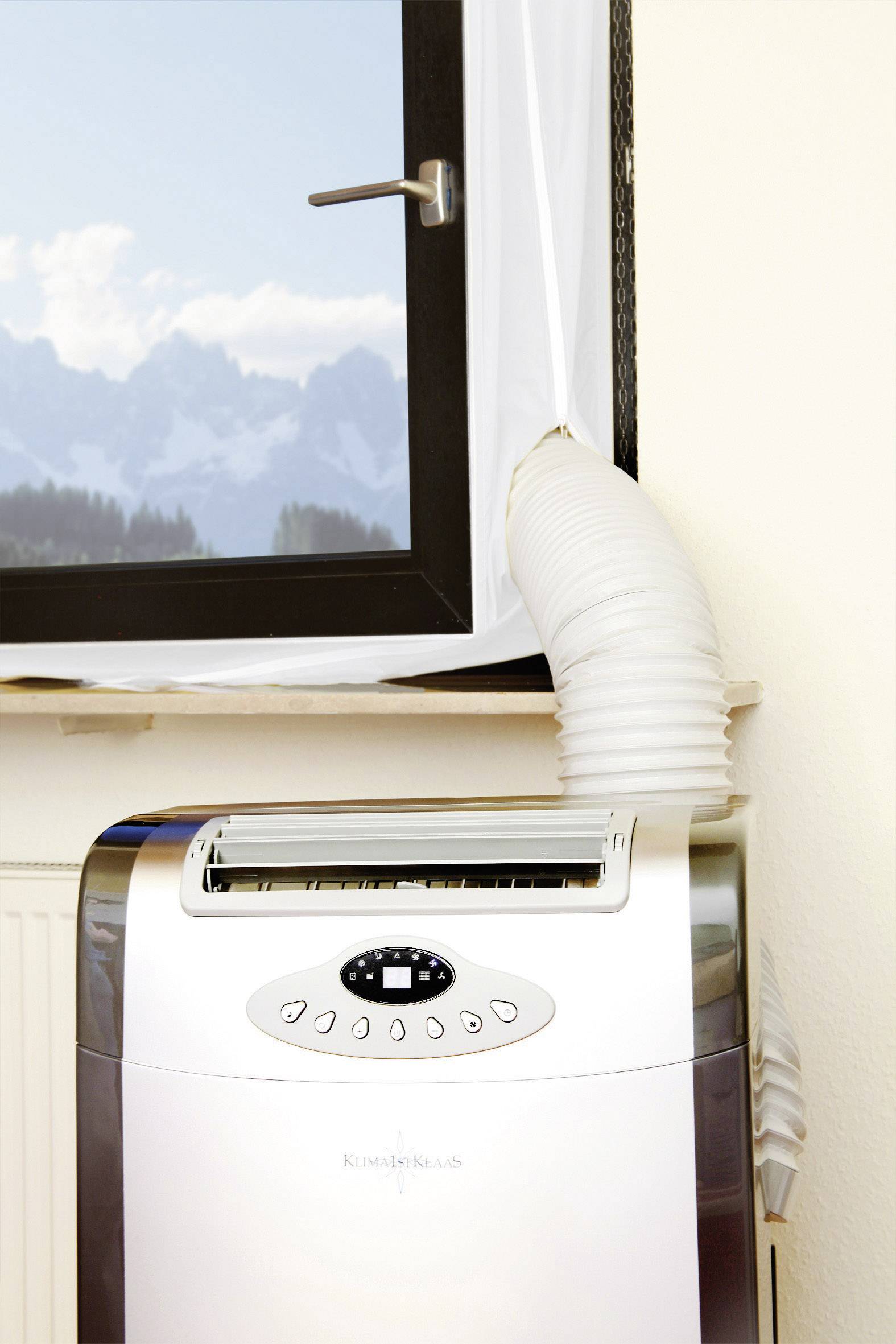 A portable air conditioning unit is positioned beneath an open window, with an extractor hose leading outside. Mountains can be seen in the background.