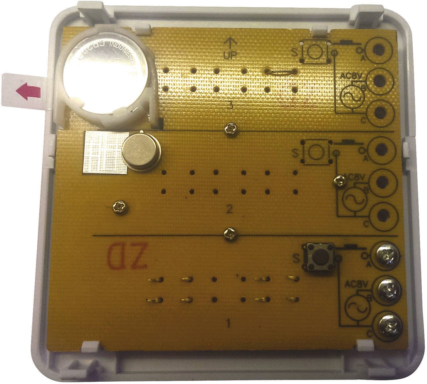 An electronic device with multiple circuit boards and buttons. A small battery is located in the top left corner.