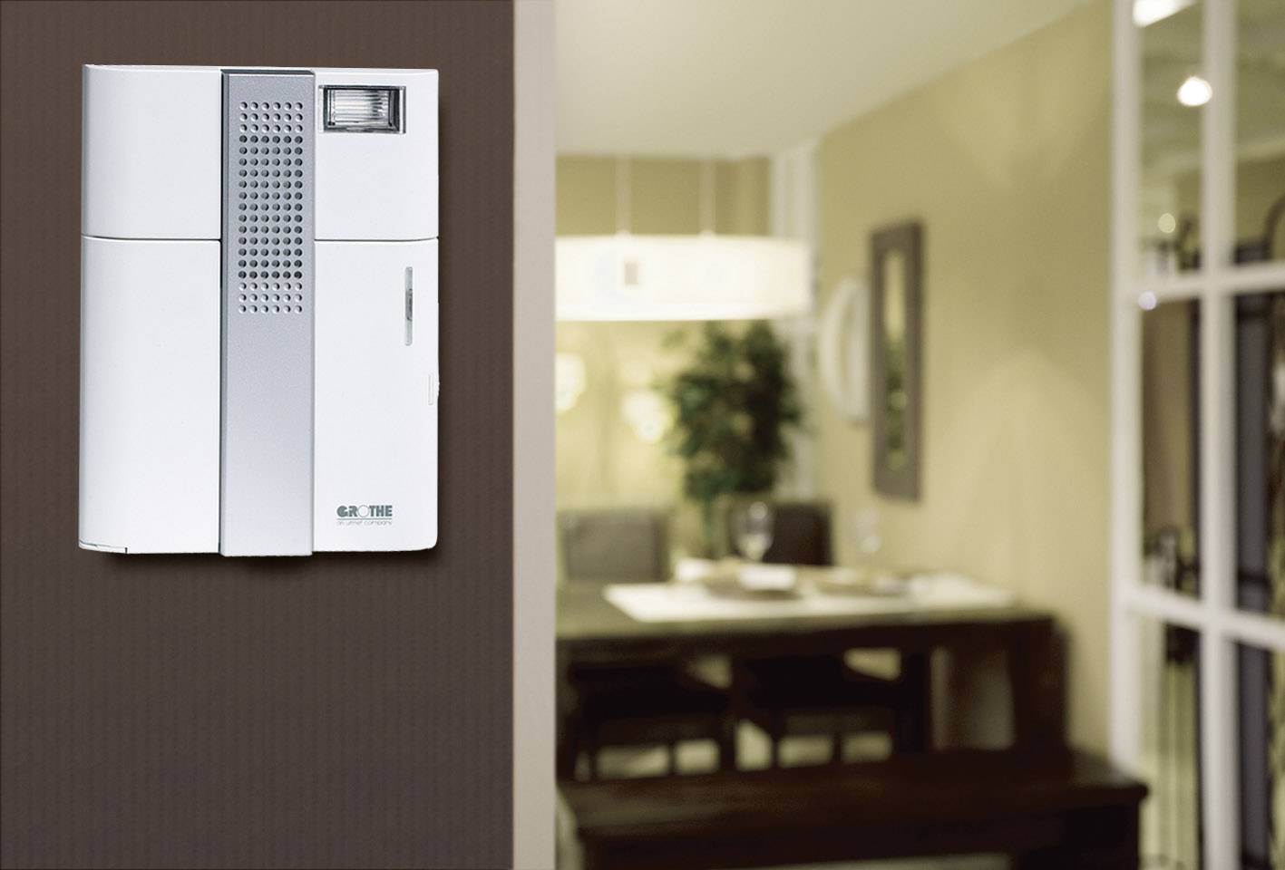 Intercom mounted on a wall, with a blurred view of a dining room with a table and chairs in the background.