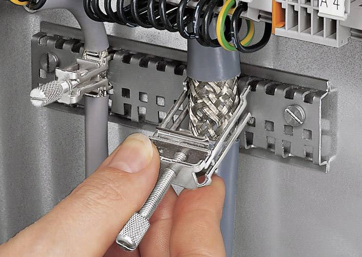 A hand secures a braided cable to a clamp on a metal surface, surrounded by several cables and cable mounts.