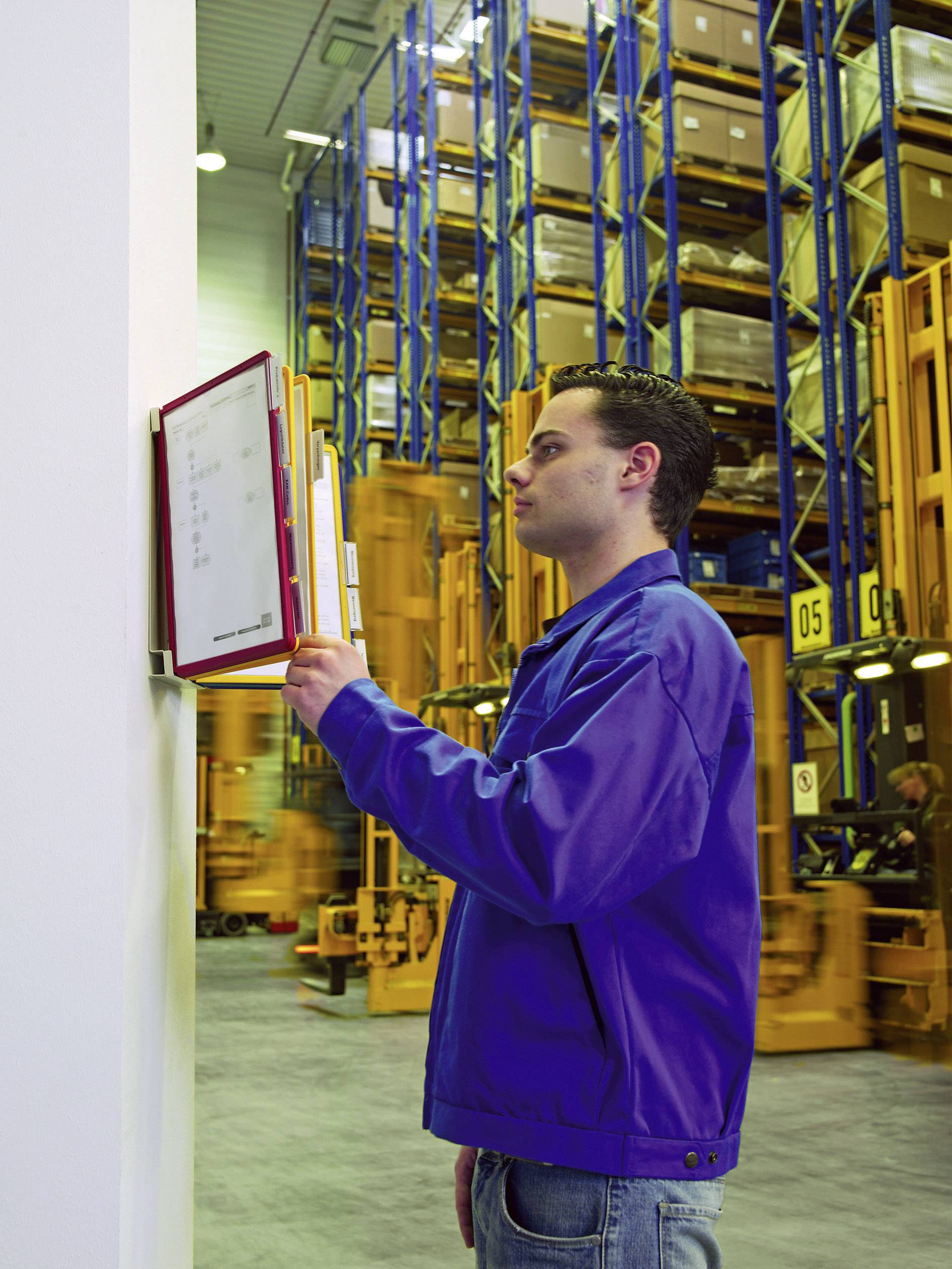 A man in a warehouse is checking documents on a clipboard. High shelves stacked with cardboard boxes are visible in the background.