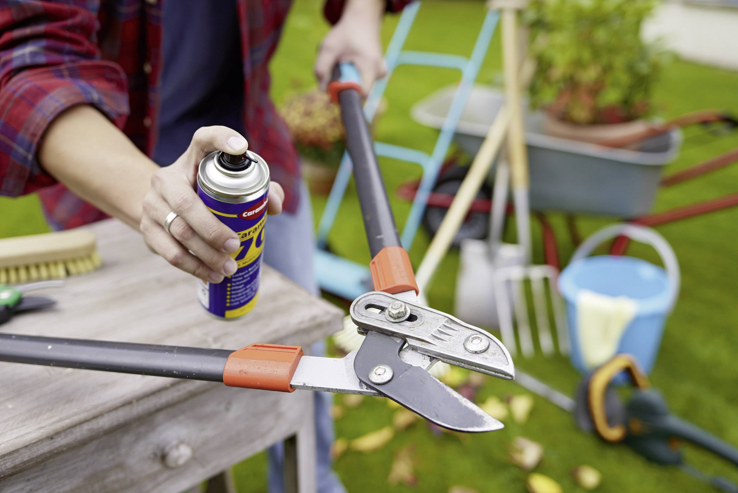 A person is spraying lubricant onto secateurs in a garden. Gardening tools and a wheelbarrow can be seen in the background.