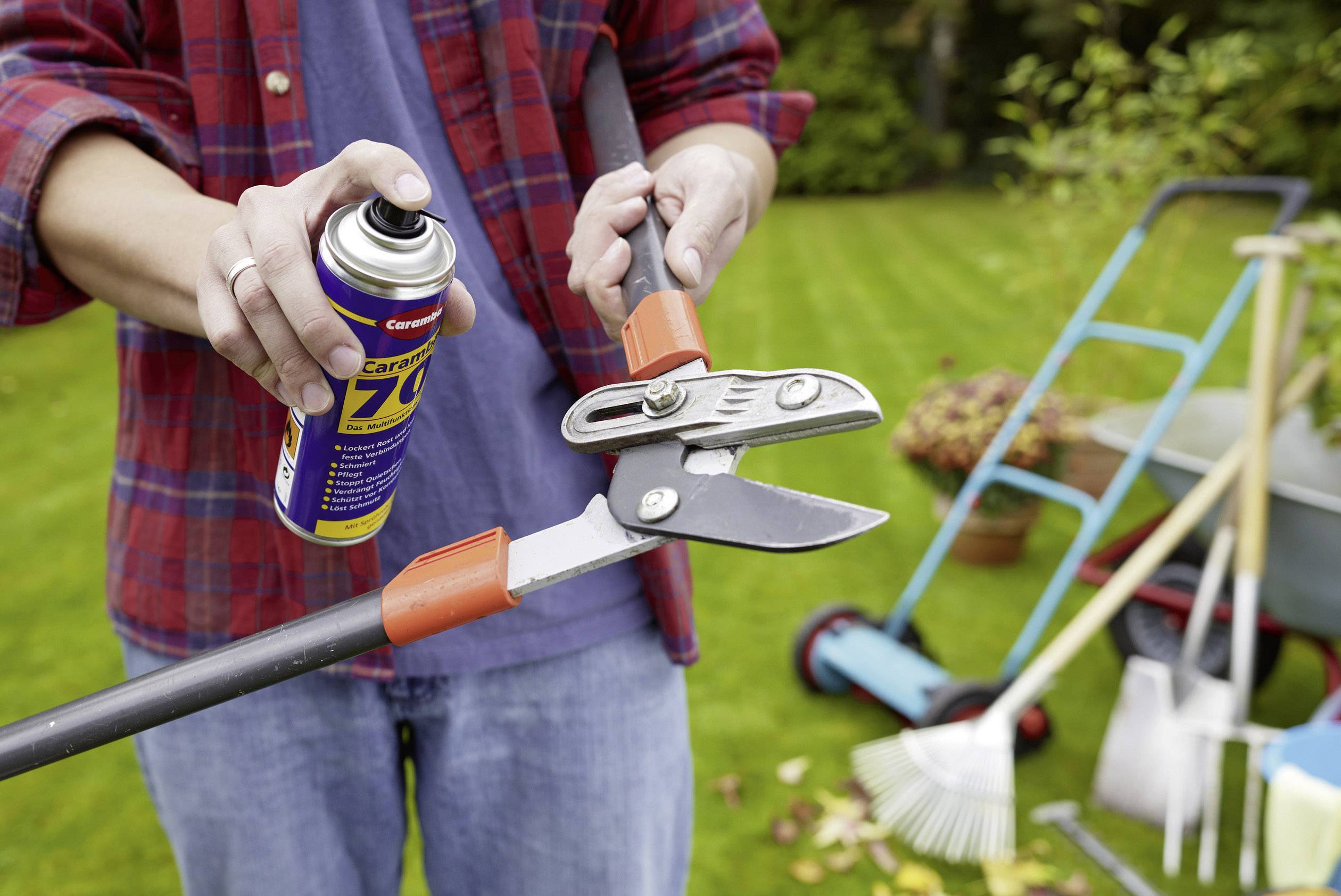 A person is lubricating garden secateurs with spray. In the background, gardening tools and a wheelbarrow can be seen.