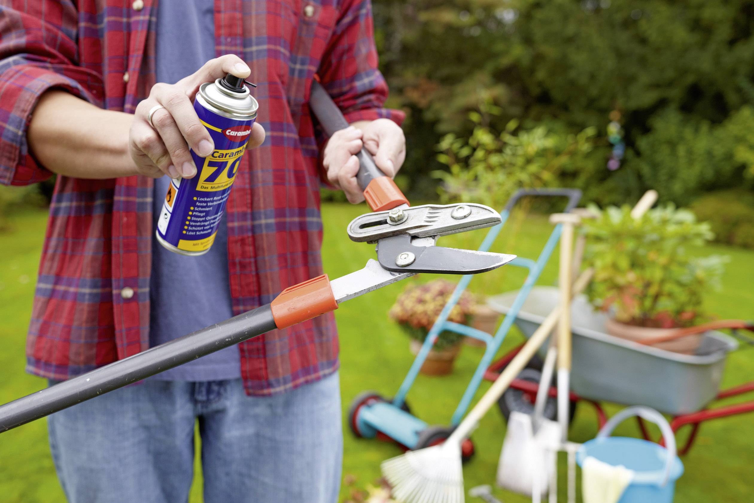 A person is spraying lubricating oil onto the joints of a pair of secateurs in the garden. Garden tools and plants can be seen in the background.