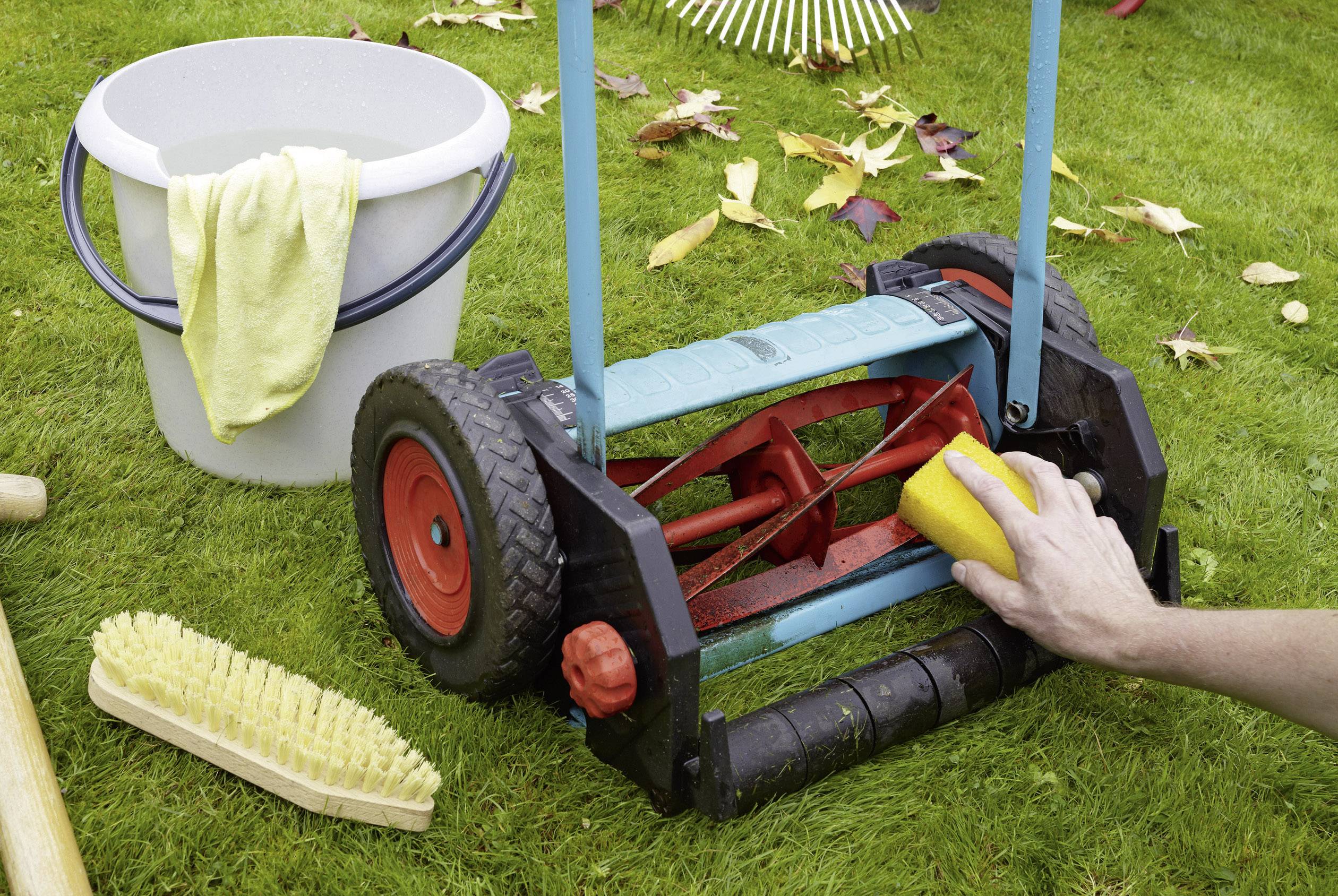 A person is cleaning a hand lawnmower with a sponge on a lawn. A bucket and a brush are visible in the background.