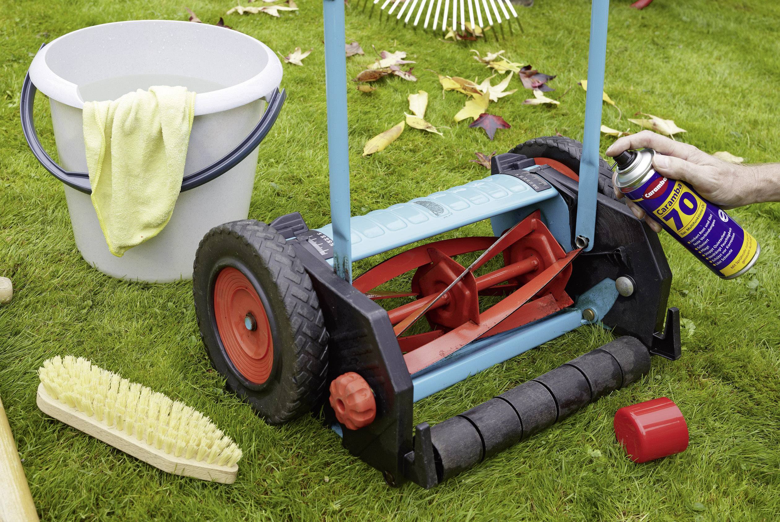 A hand is spraying oil onto the red and blue roller of a lawnmower. In the background, a bucket, a cloth, leaves, and a brush can be seen.