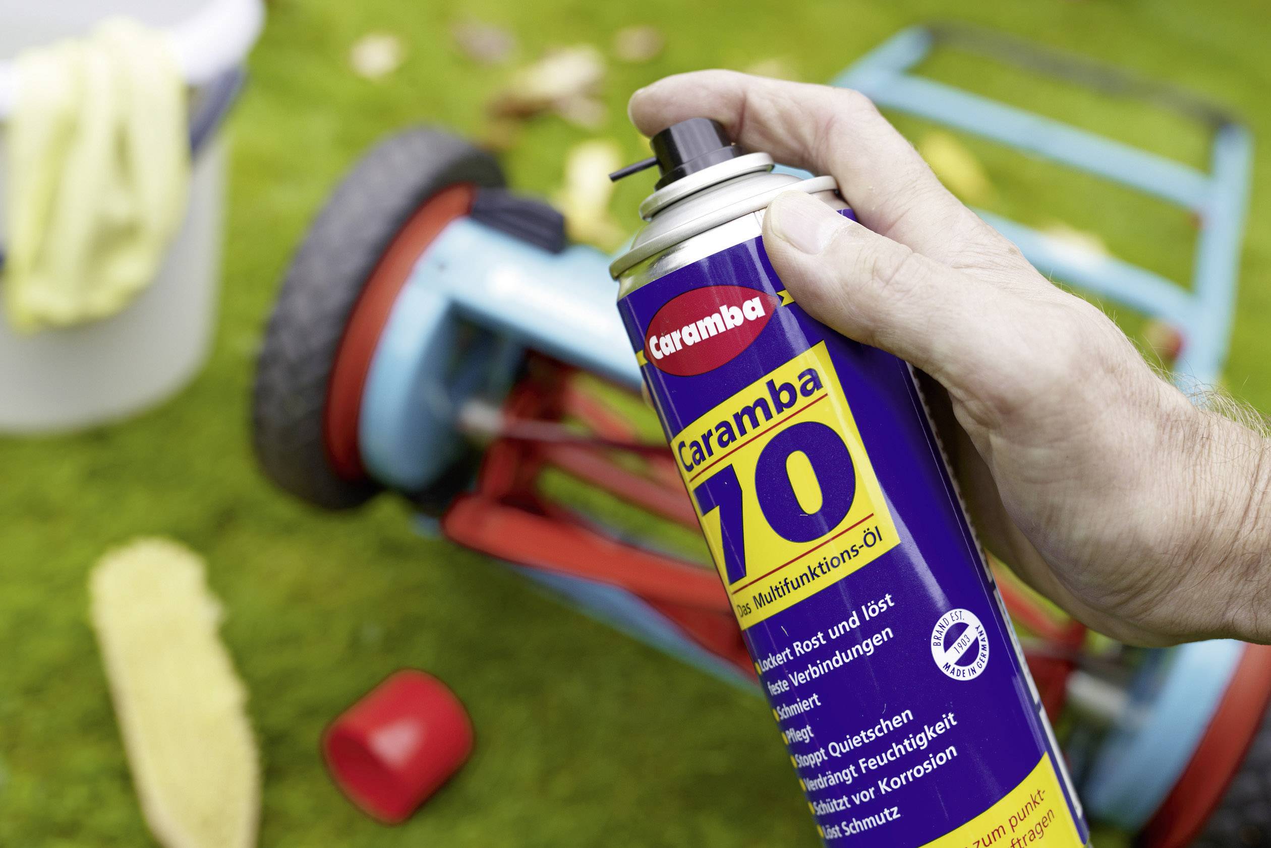 A person is holding a tin of Caramba multi-purpose oil. In the background, an overturned lawnmower lies on a green lawn.