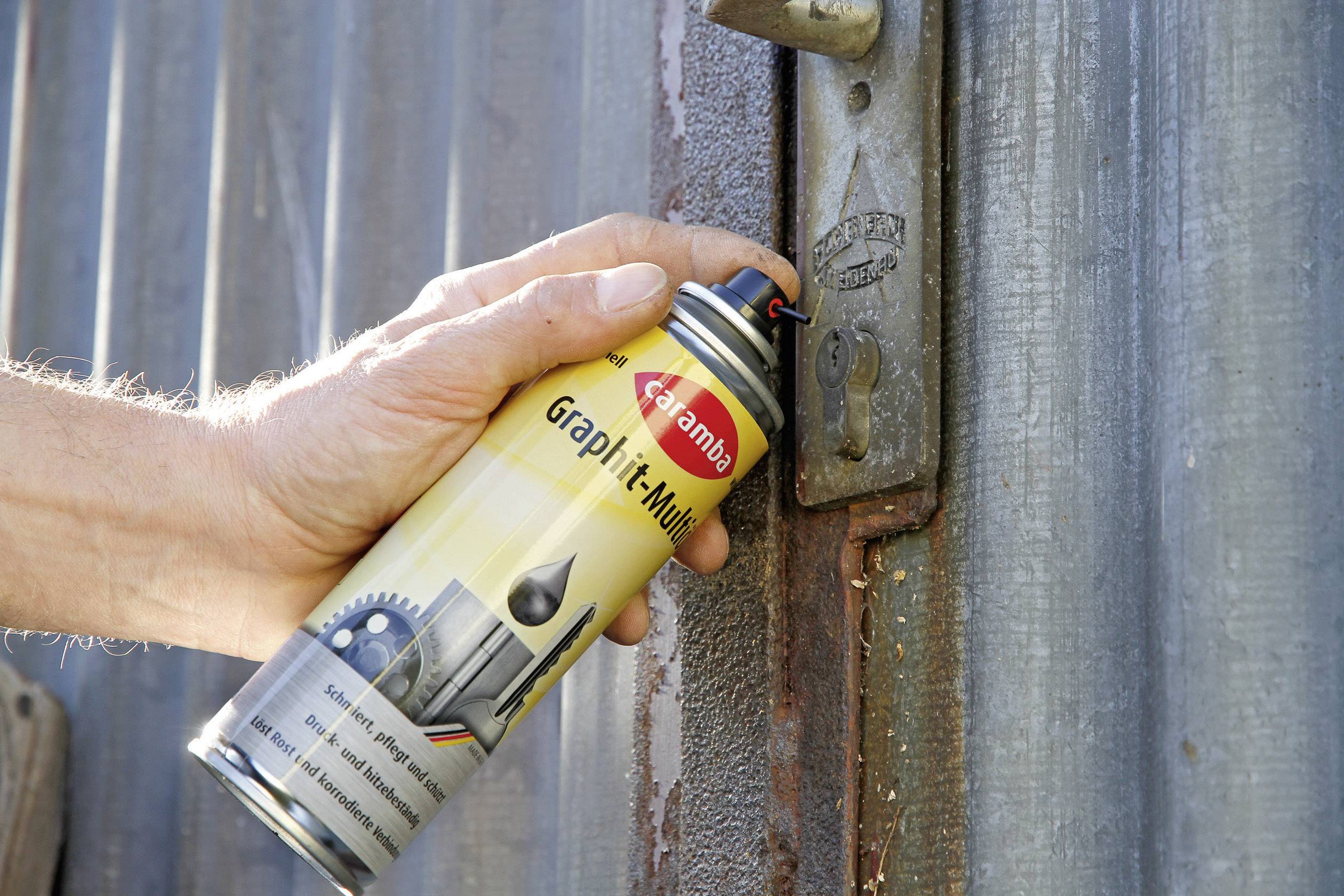 A hand sprays lubricant from a can onto a rusty door lock on a metal gate to lubricate and protect it from rust.