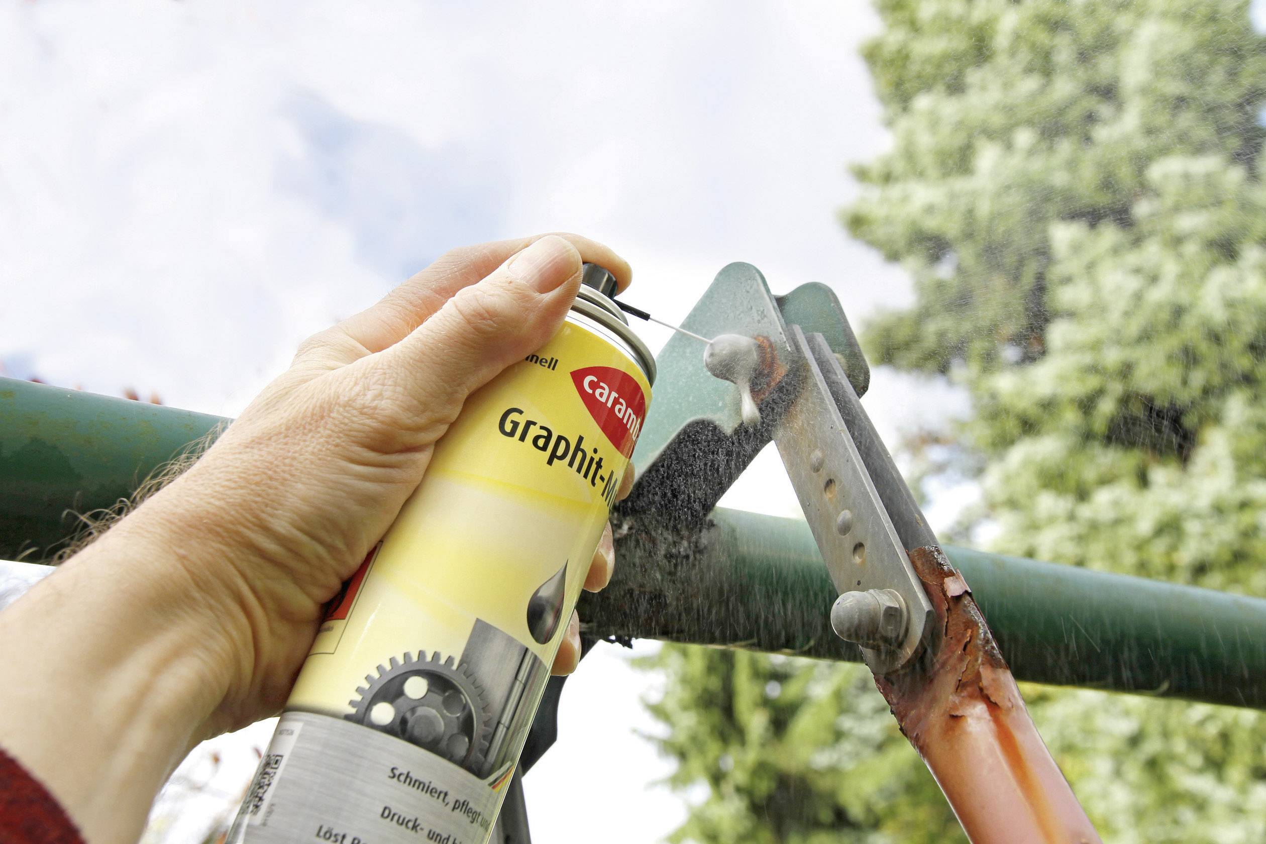 A person sprays lubricant onto pruning shears to maintain them. Trees can be seen in the background.
