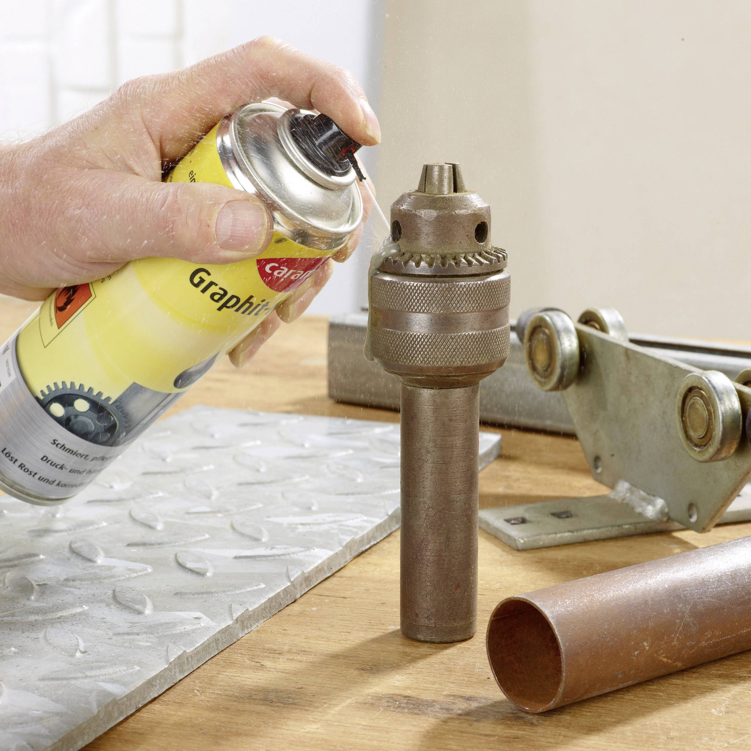 A hand sprays graphite spray onto a metal drill bit and a metal plate. Beside it, a pipe lies on a workbench.