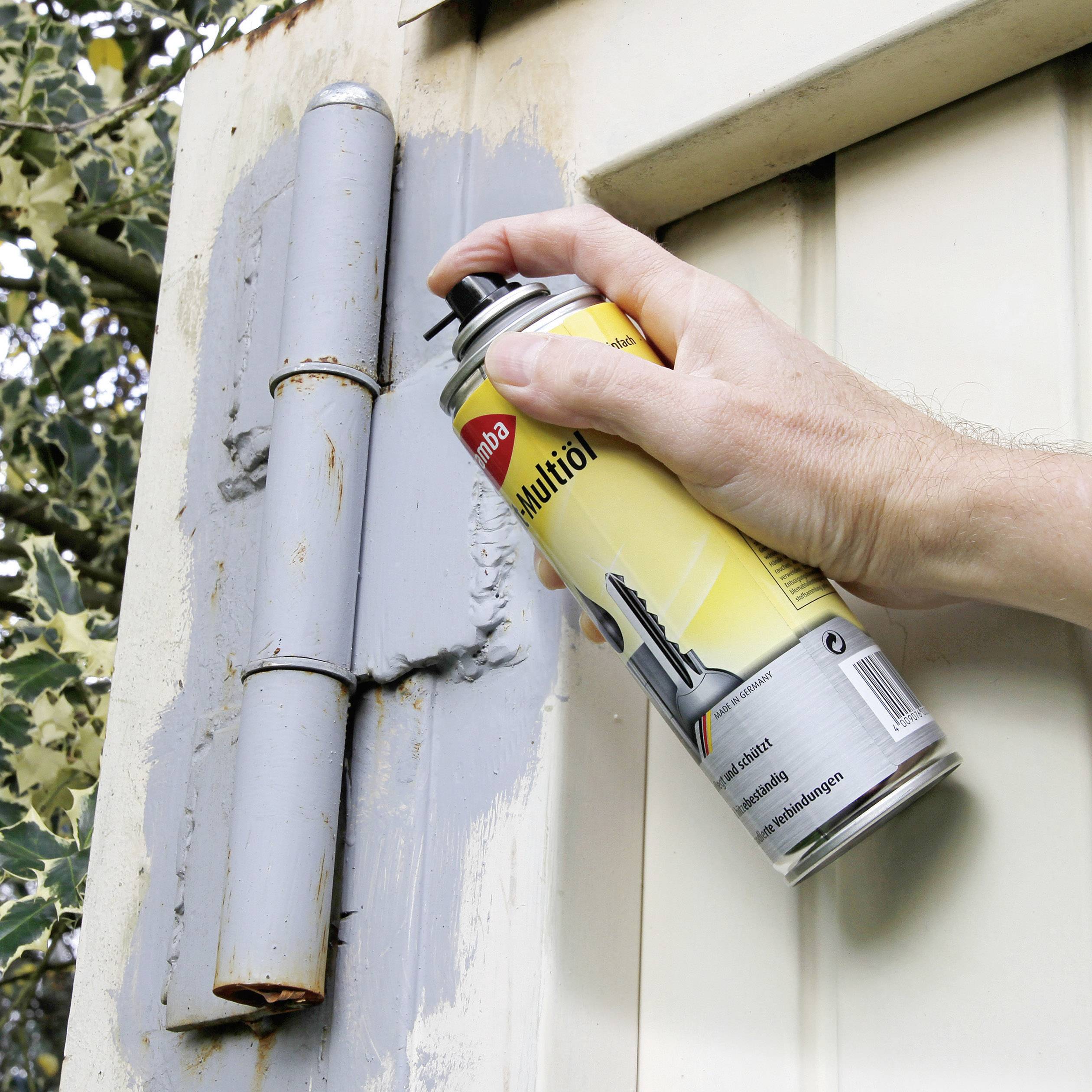 A hand sprays lubricant onto a rusty door hinge of a metal gate to ease movement and reduce corrosion.