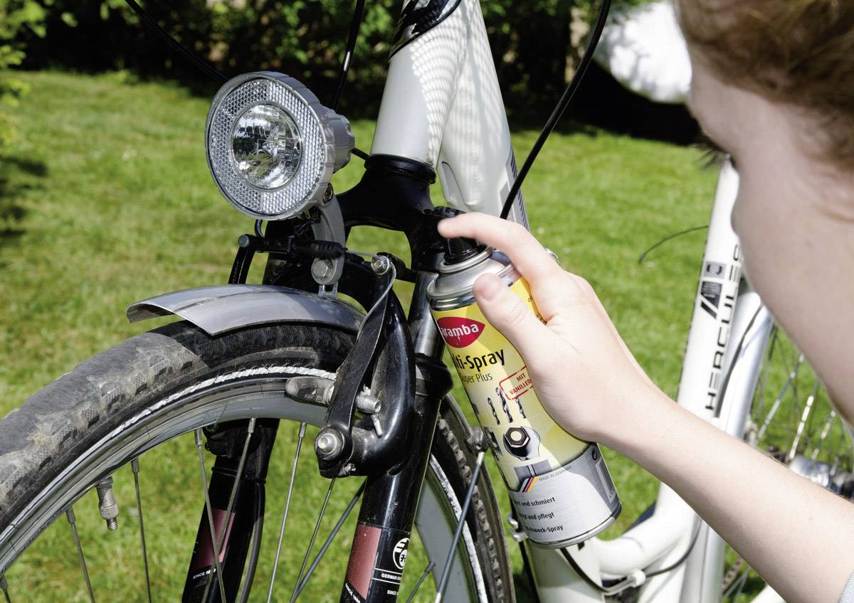 A person sprays a bicycle fork with rust protection; focus on the front light and rim of the black bicycle against a green background.
