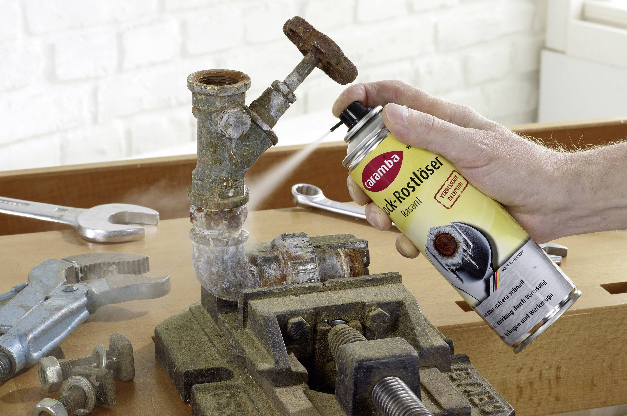 A person is spraying rust remover onto a rusted pipe connection in a workshop. Tools and a spanner are lying in the background.