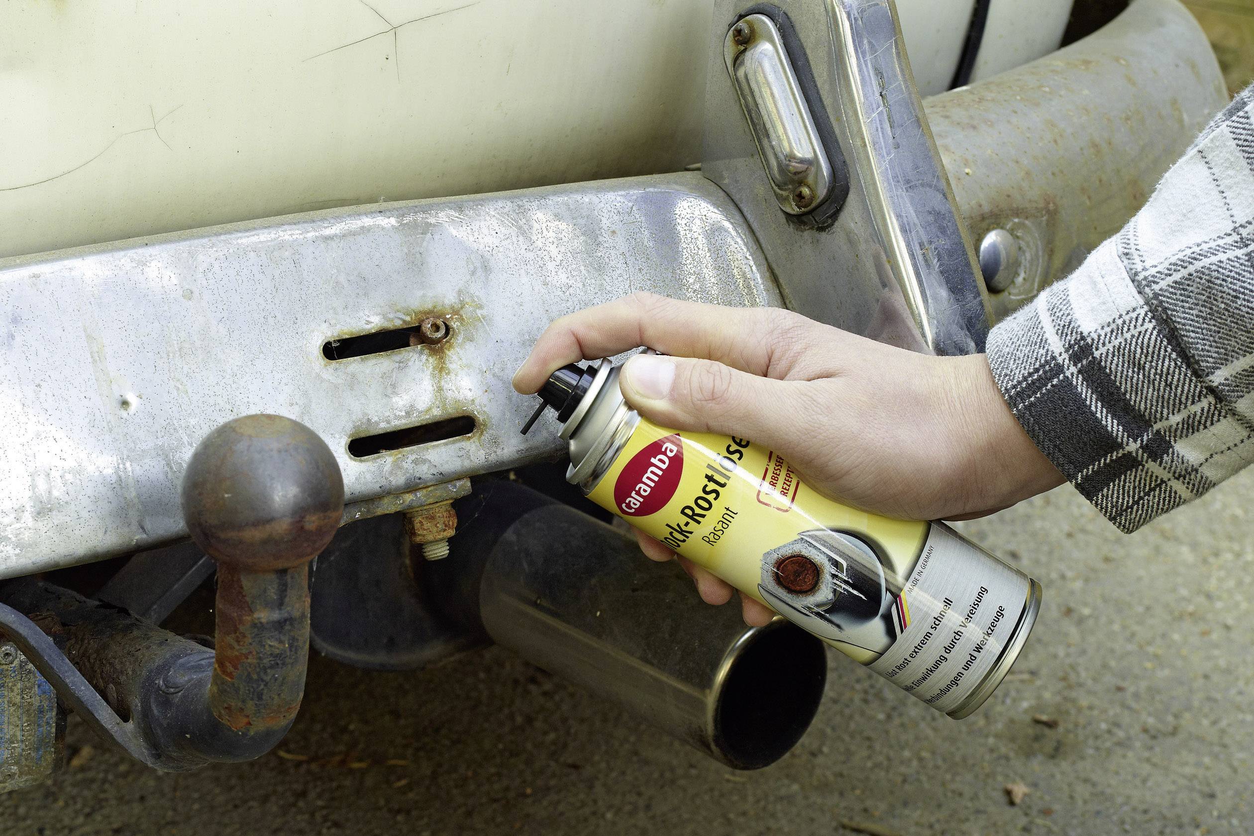 A person is spraying rust protection spray onto a rusty car bumper to prevent corrosion.
