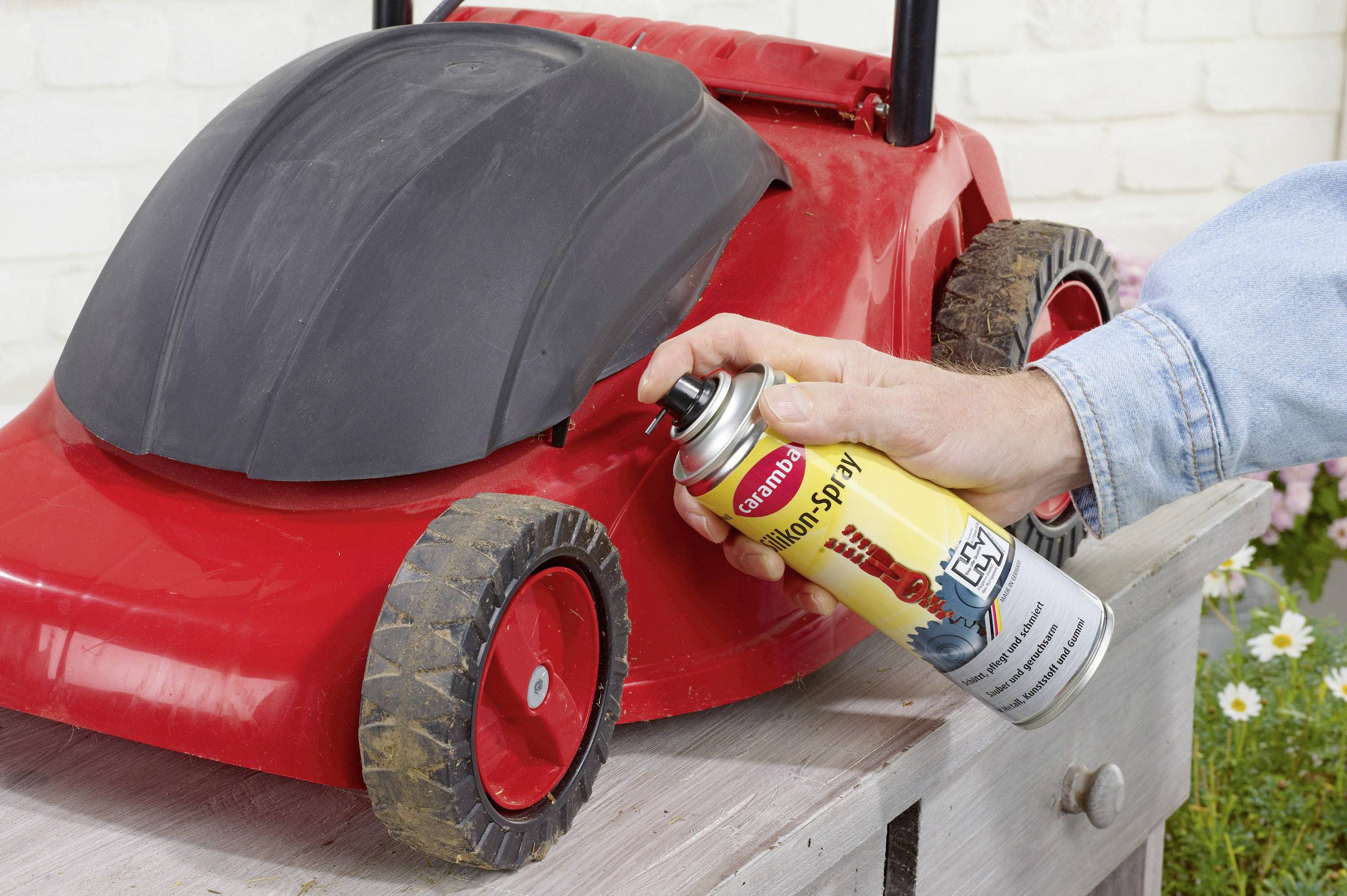 A person is spraying a red lawnmower with silicone spray for maintenance and care on a wooden table outdoors.
