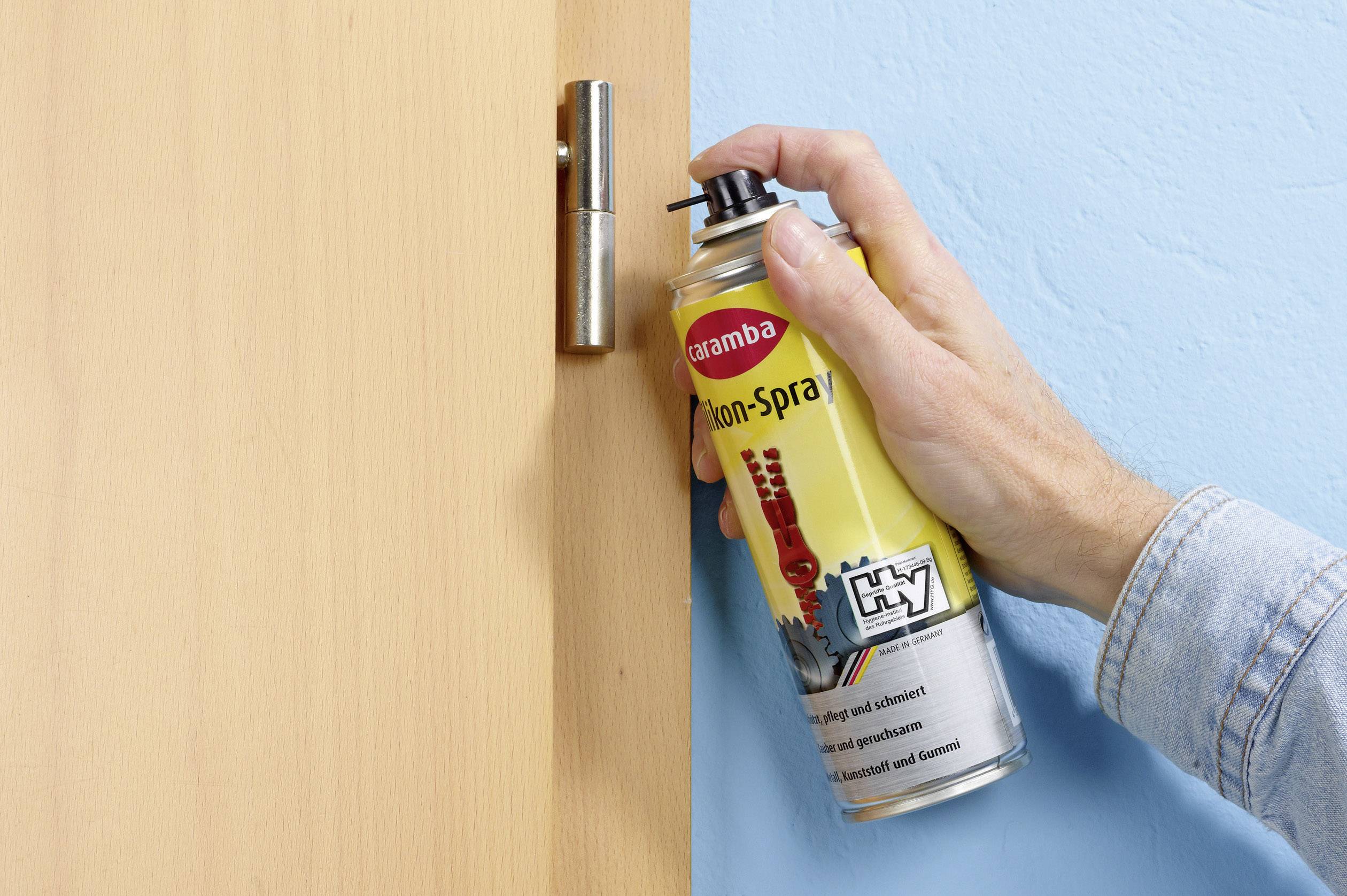 A hand sprays lubricant on a door hinge to maintain it and eliminate any potential squeaking.