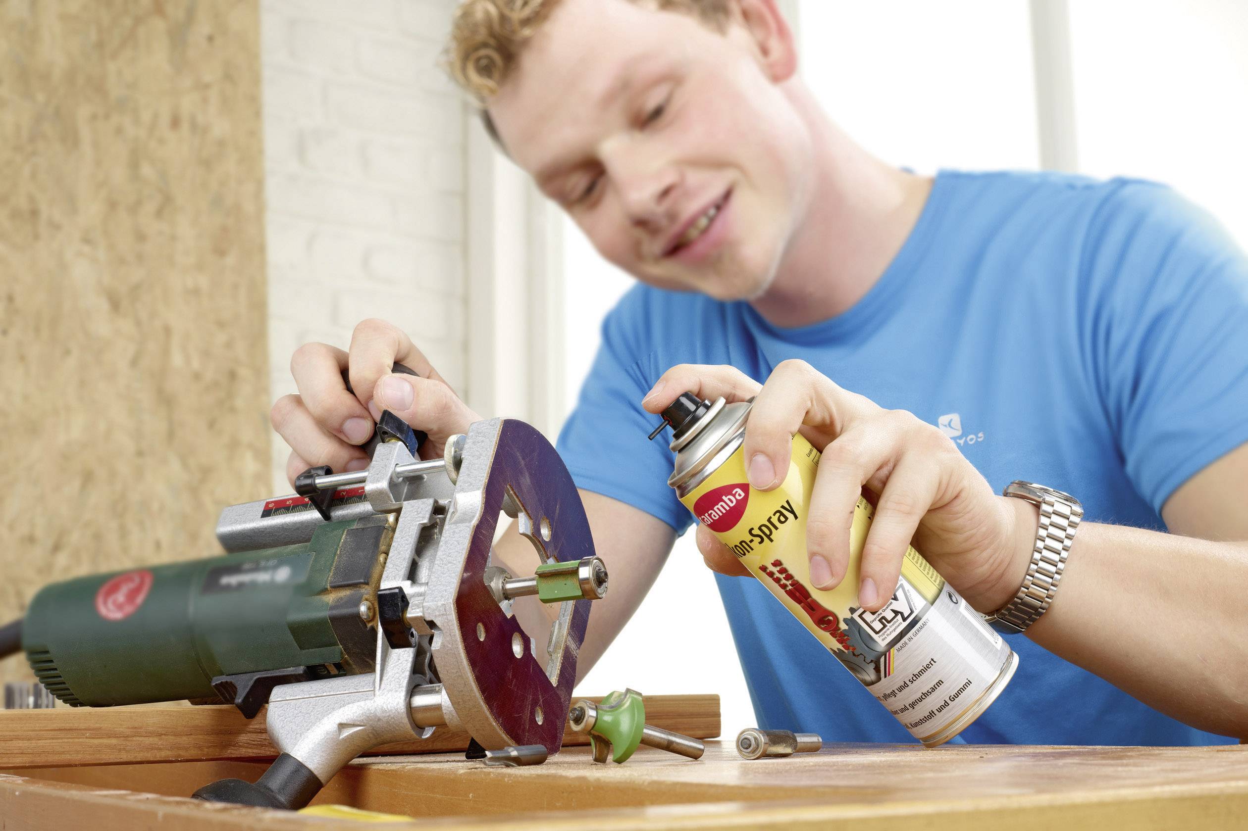 A man is lubricating an electric tool in a workshop. He is wearing a blue T-shirt and using a WD-40 spray for maintenance.