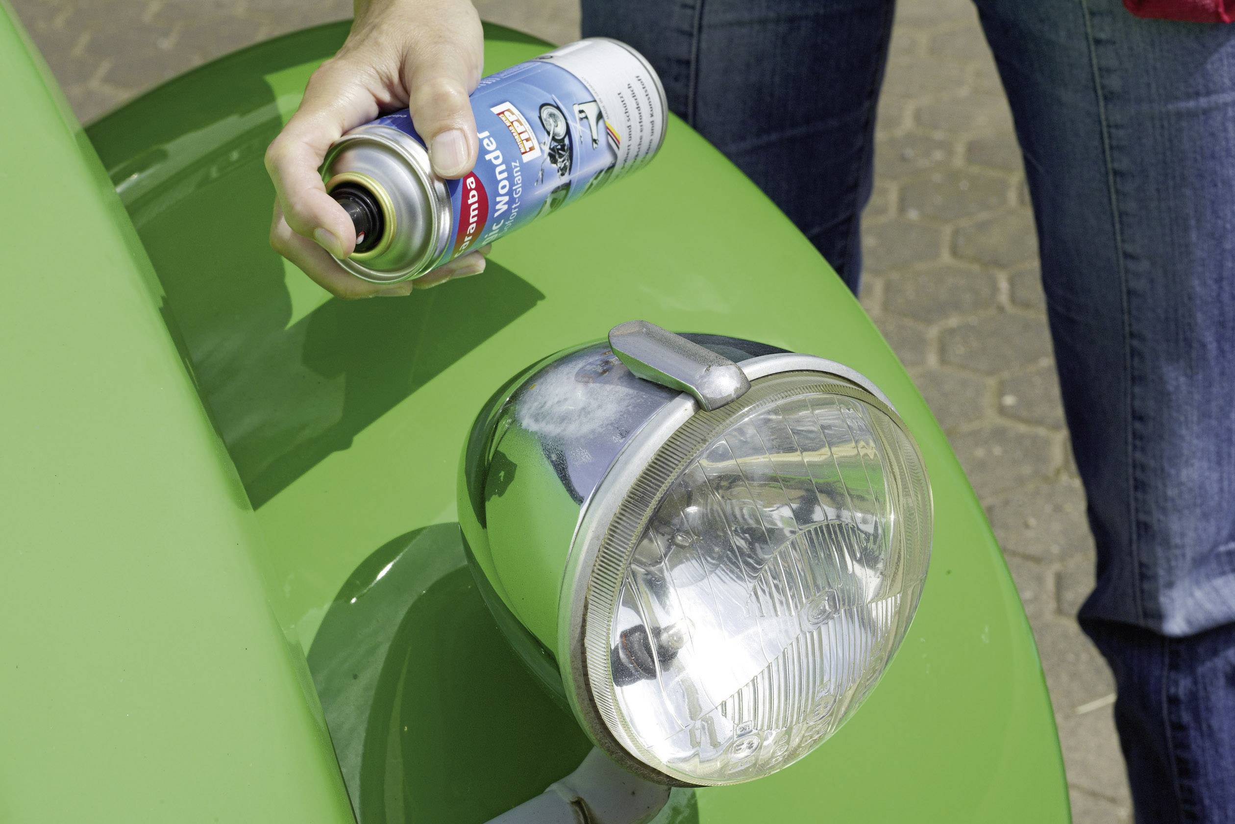A person is holding a spray can and spraying onto the bonnet of a green vehicle, near a round headlight.