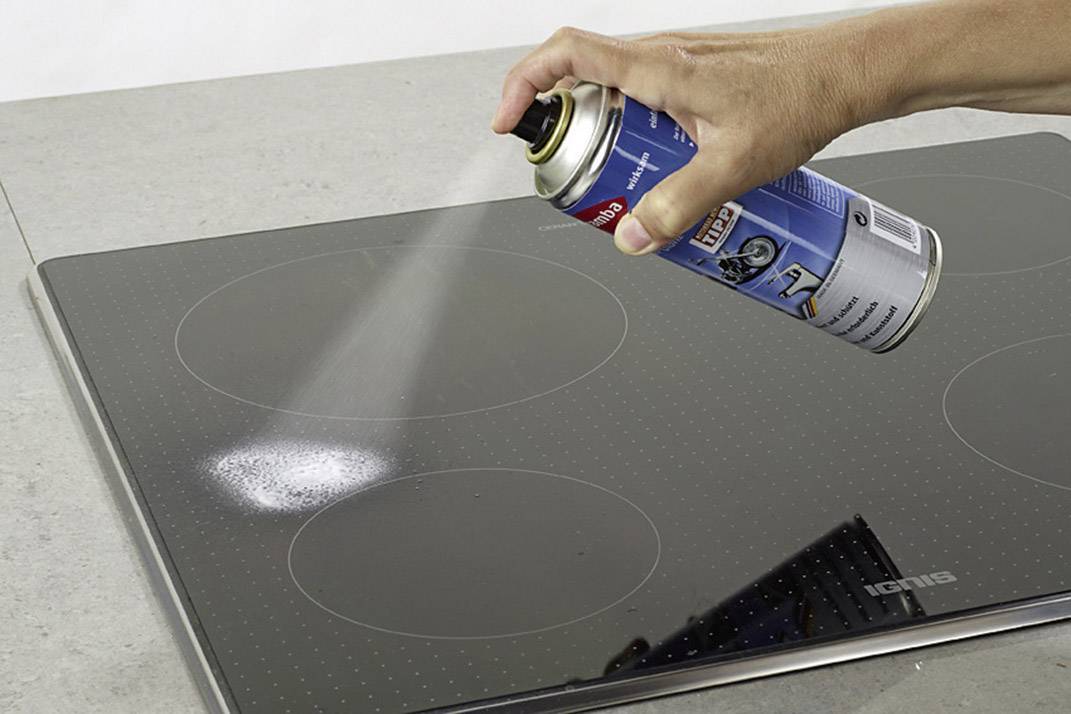 A hand sprays cleaner from an aerosol can onto a black ceramic hob. Four cooking zones are visible.