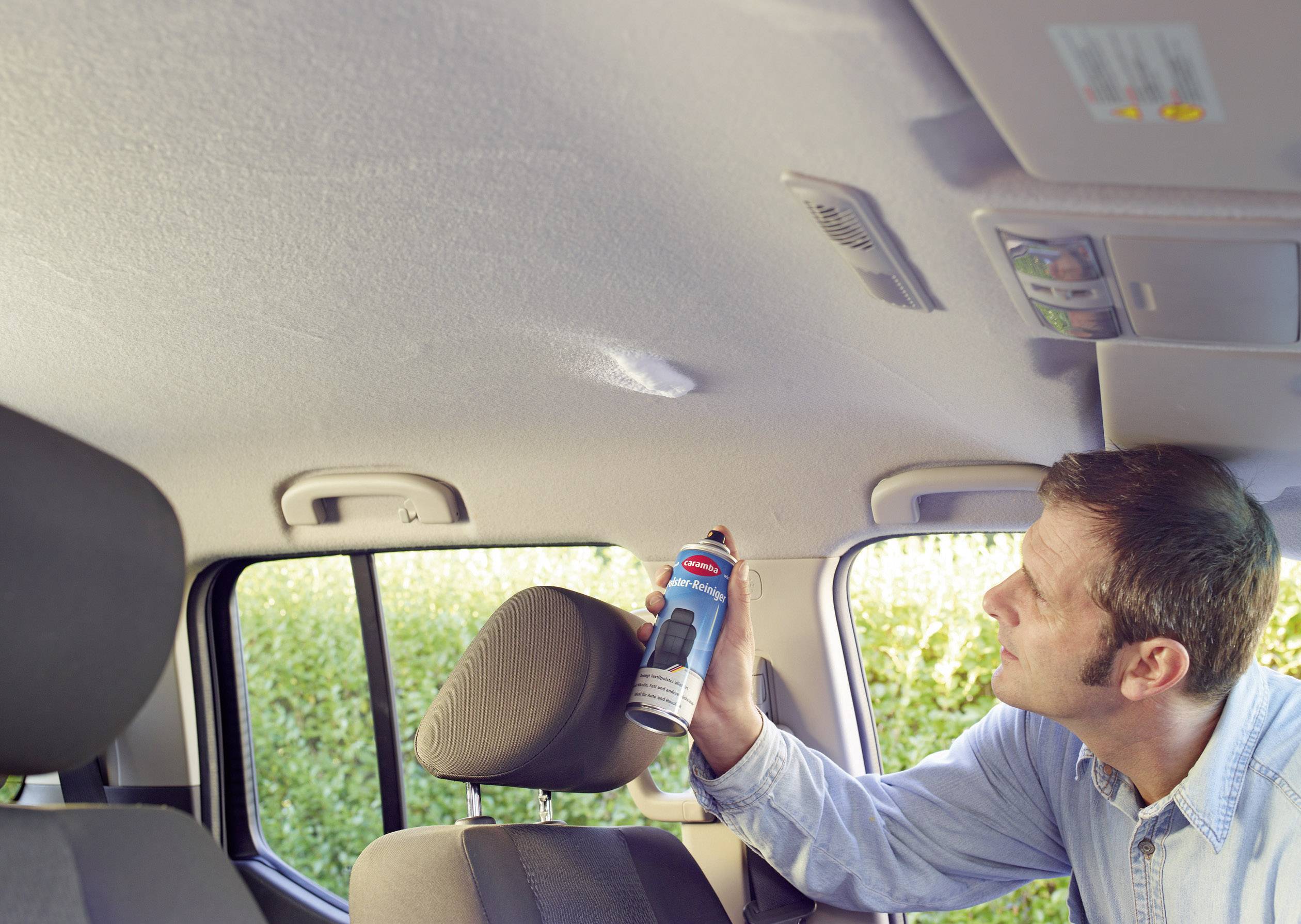 A man is using a spray can to clean the car's interior roof lining. He is focusing on a specific area.