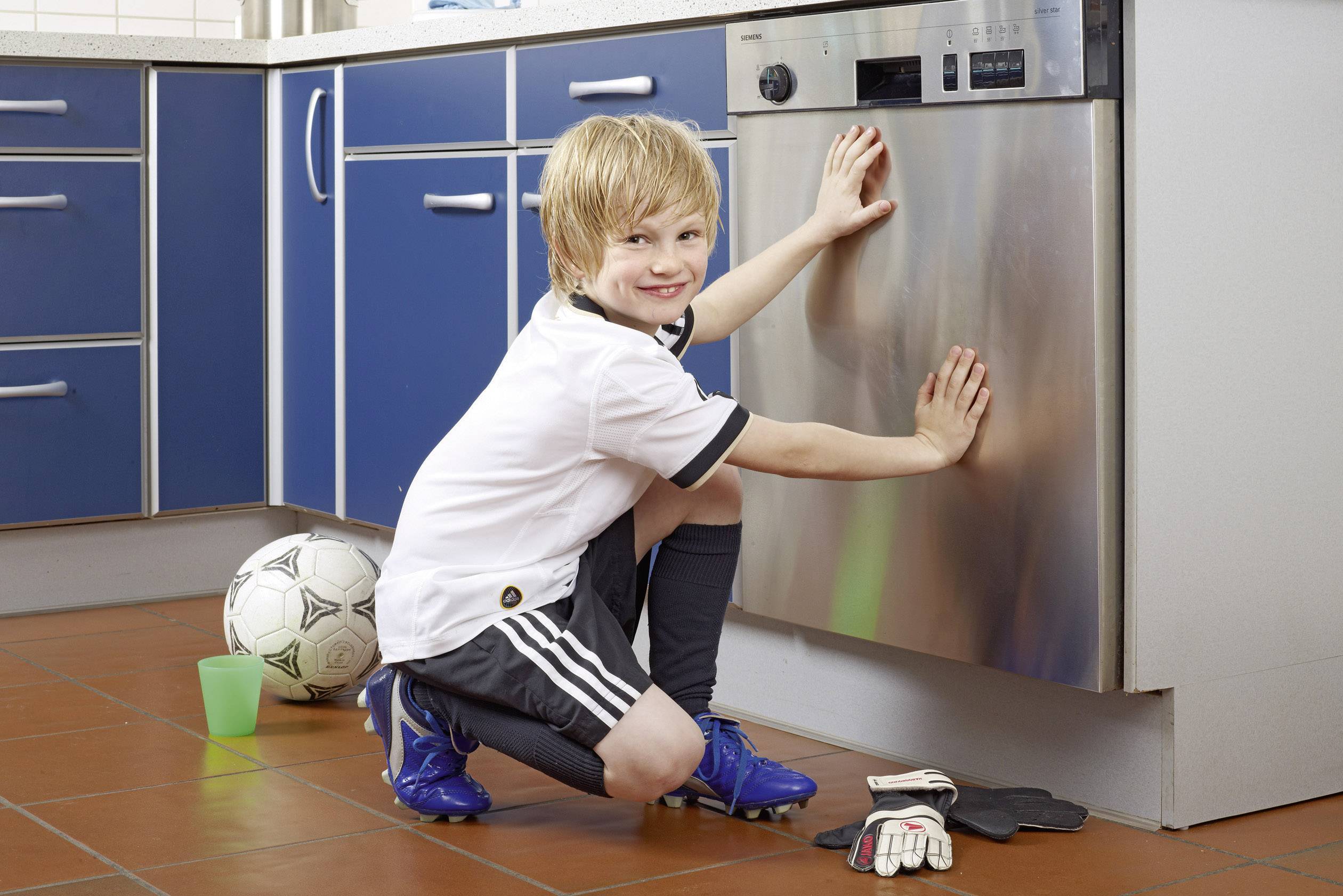 A child in football kit is cleaning the door of a dishwasher in a kitchen with blue cupboards. A football and gloves are lying on the floor.